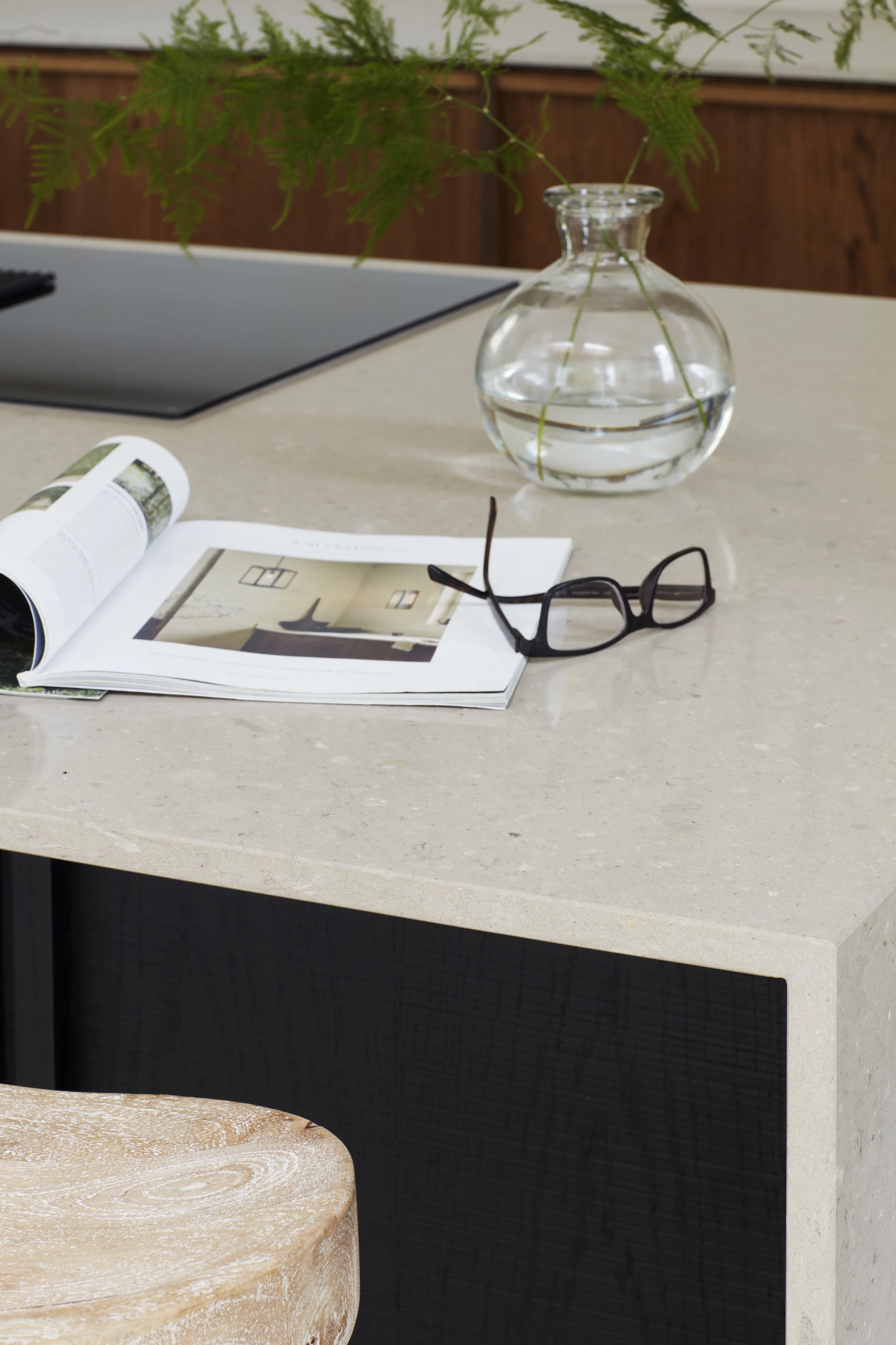 A view of a beige stone tabletop with an open magazine, a pair of black eyeglasses, and a glass vase with water and green fern leaves inside.