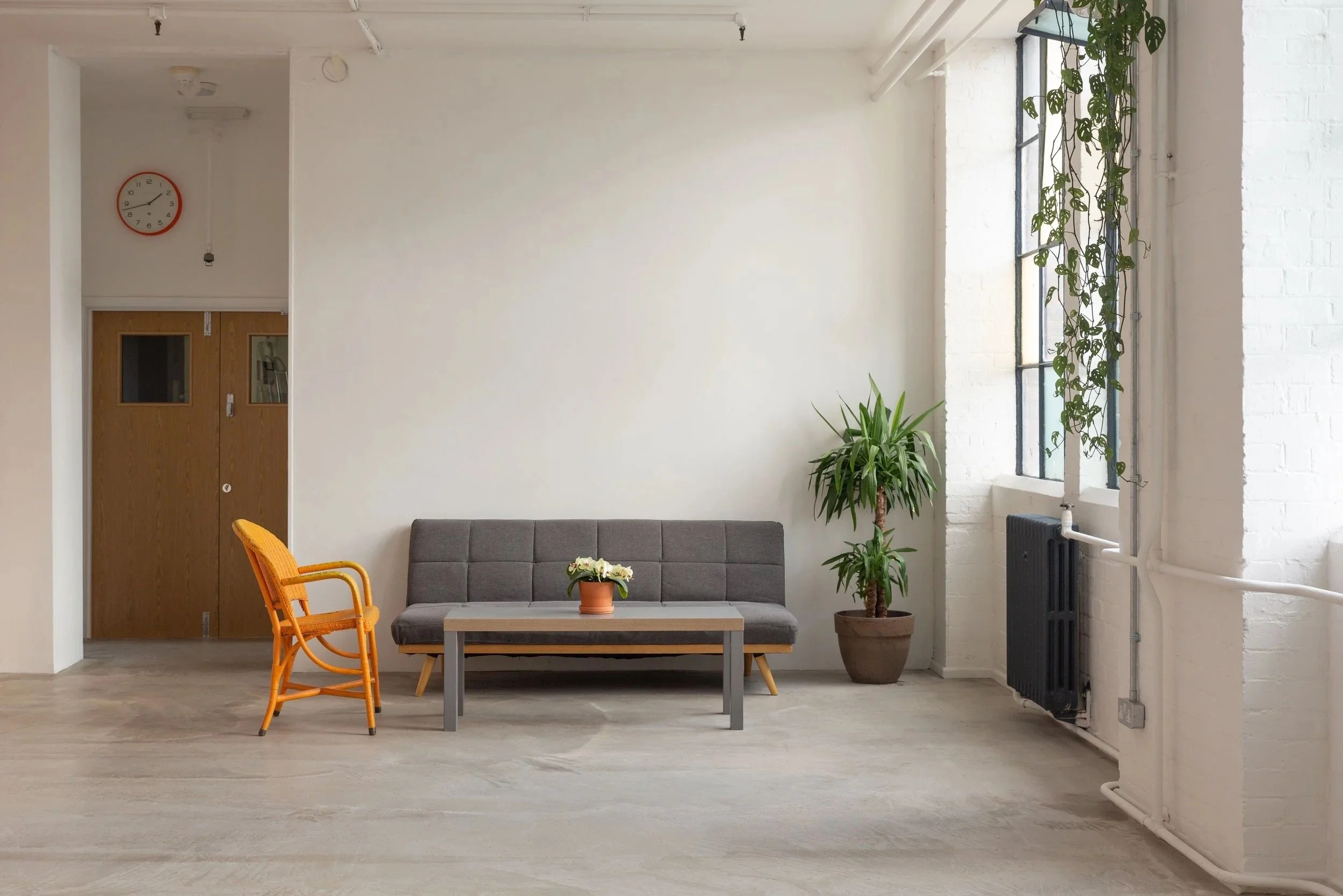Interior of a minimalistic living room with a gray sofa, a small coffee table with a potted plant, an orange rattan chair, a large potted plant, and a window with hanging plants, white brick walls, and a wooden door with a clock above.