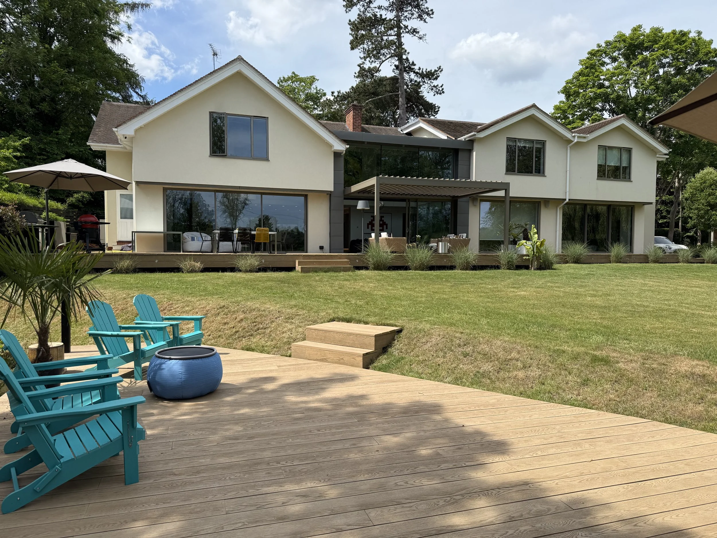 Large modern house with a deck, outdoor seating, and lawn in a suburban setting during daytime.