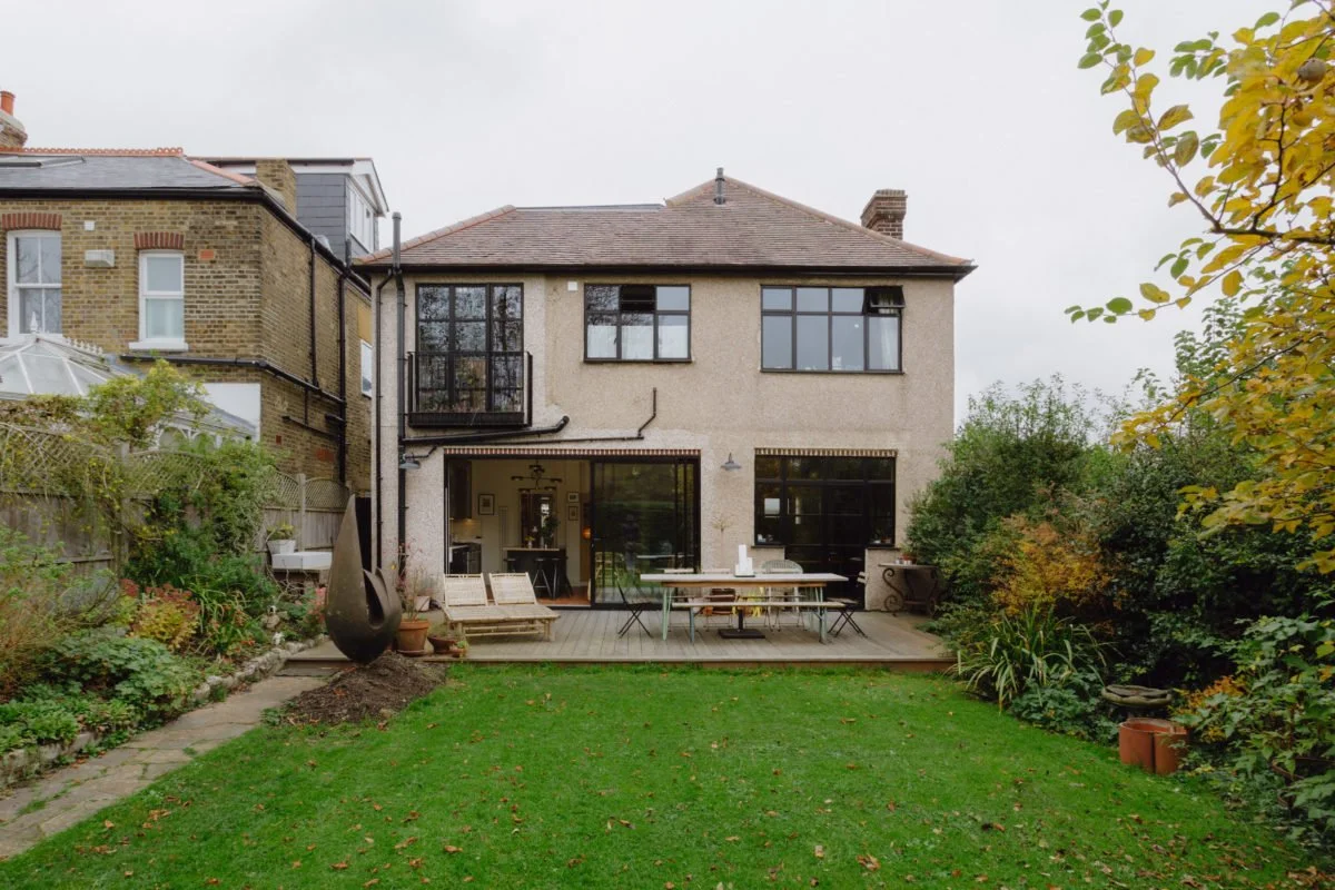 Rear view of a two-story house with a garden and outdoor seating area, featuring wooden decking and lush greenery.