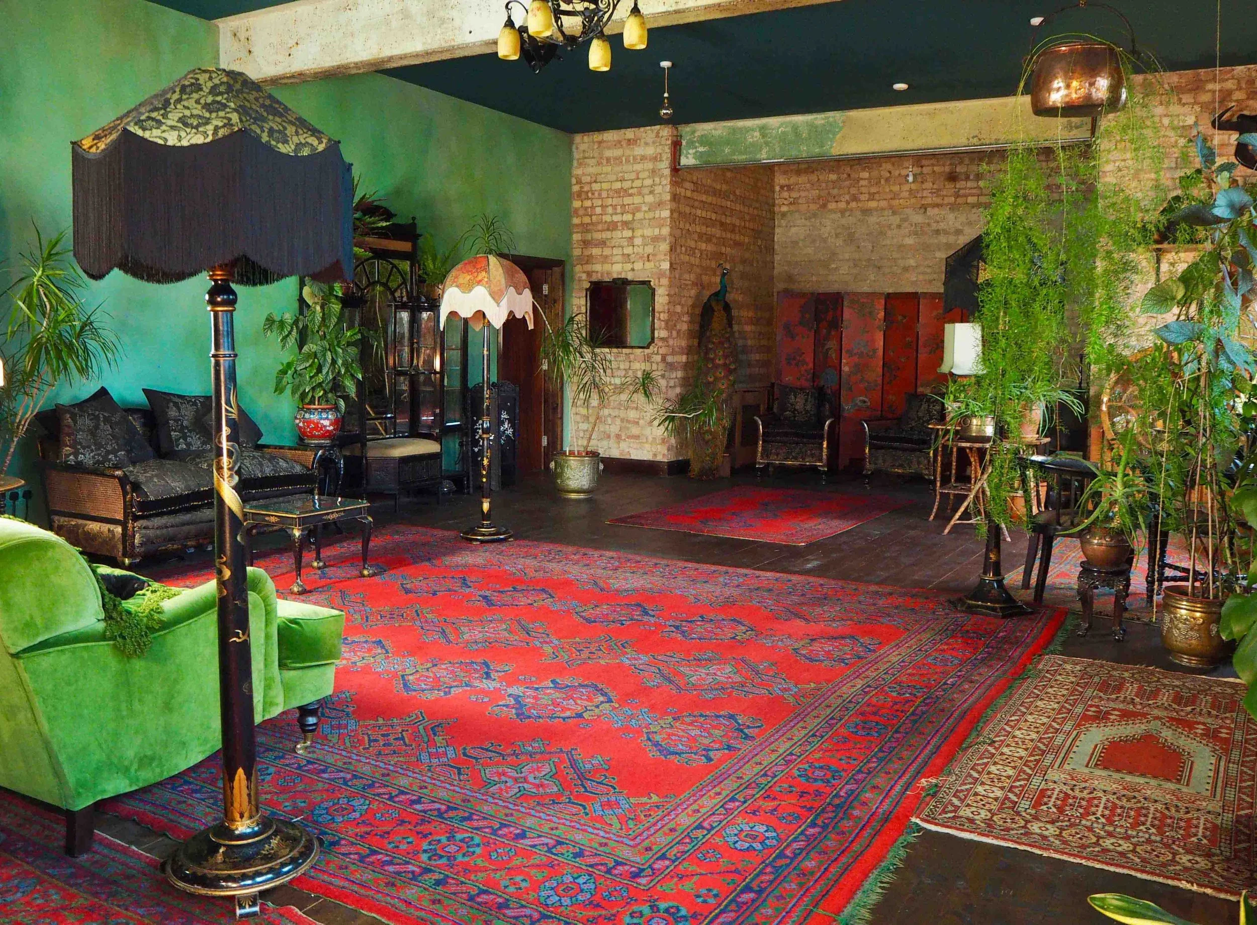 A spacious living room with vintage furniture, colorful rugs, and many green plants. The walls are painted green and exposed brick, with various lamps and framed decorations.