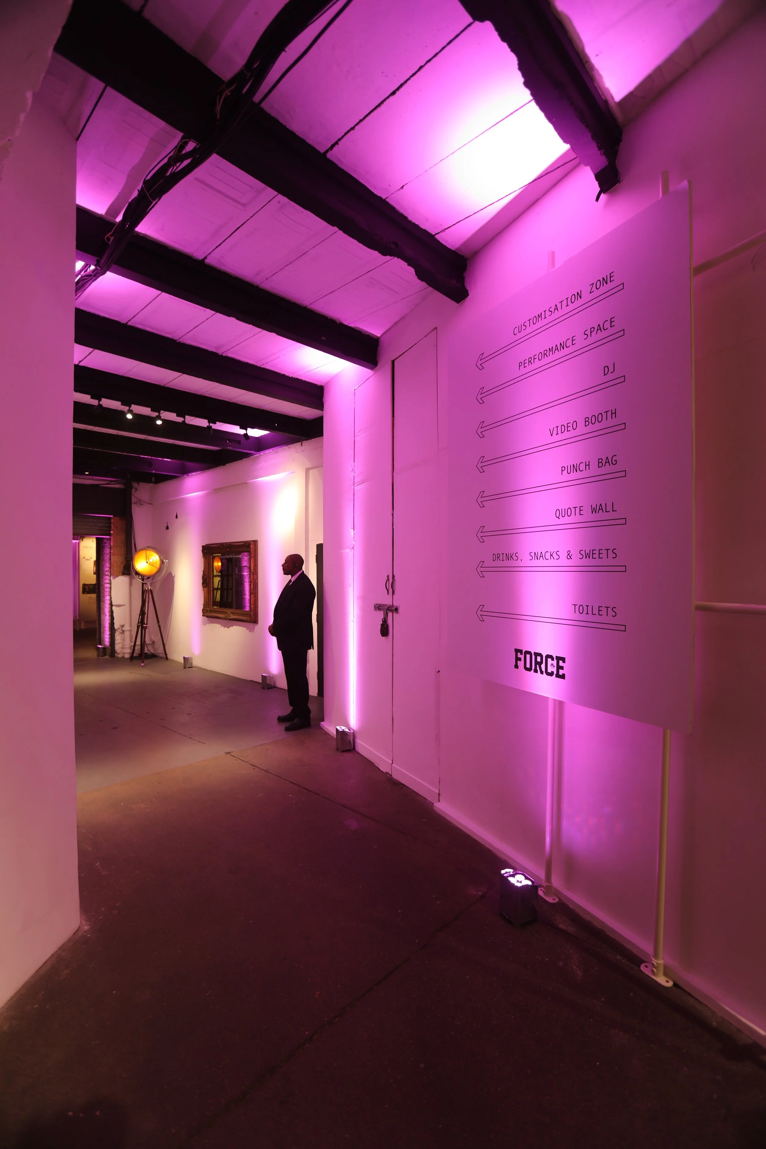 A hallway with purple lighting and a man in a suit standing near a wall. There is a large sign with directions to various areas, including 'Performance Space,' 'DJ,' 'Video Booth,' 'Punch Bag,' 'Quote Wall,' and 'Toilets,' with an arrow pointing left