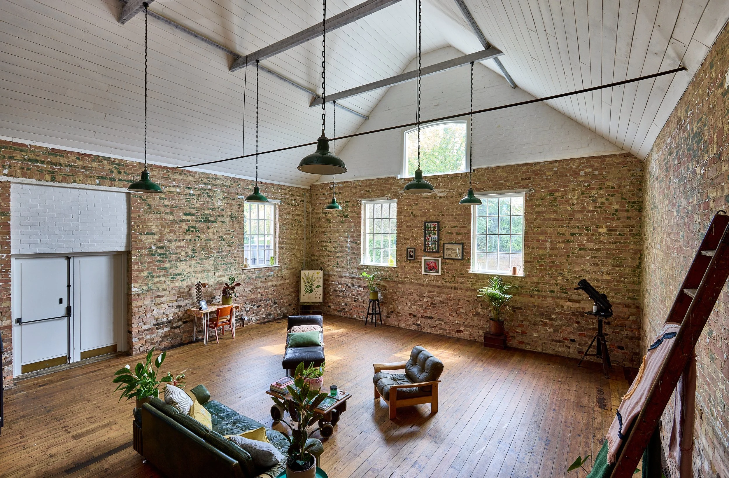 Interior of a spacious loft with exposed brick walls, a vaulted ceiling painted white, large windows, hardwood floors, hanging green pendant lights, and furniture including sofas, chairs, and a coffee table, decorated with plants and framed artwork.