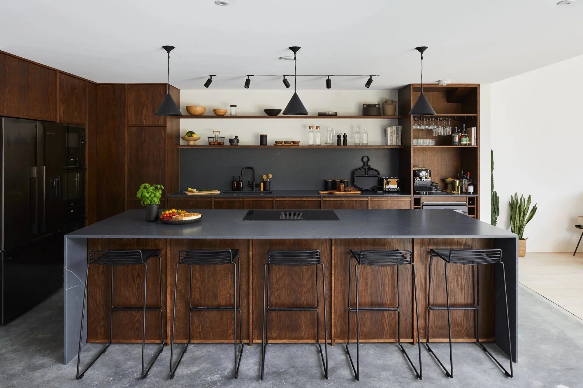Modern kitchen with dark wood cabinets, black countertops, and a large central island with five black bar stools. Open shelving with bowls, glassware, and kitchen items, and a potted basil plant on the island.