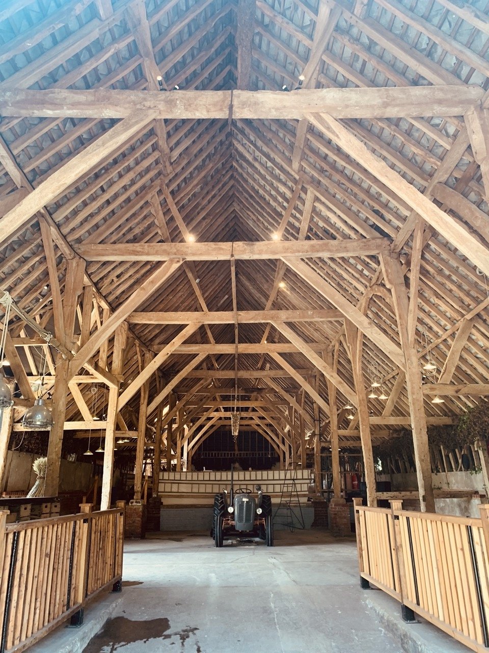 The interior of a large, open wooden barn with a tractor in the center, wooden railings on sides, exposed beams, and a high, peaked roof with lighting fixtures.