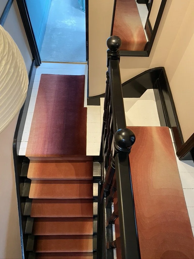 A top-down view of a staircase with a dark wooden banister and wooden steps, next to a black-framed mirror on the wall. There are red and beige rugs on the steps and landing, leading to a doorway with a blue mat outside.