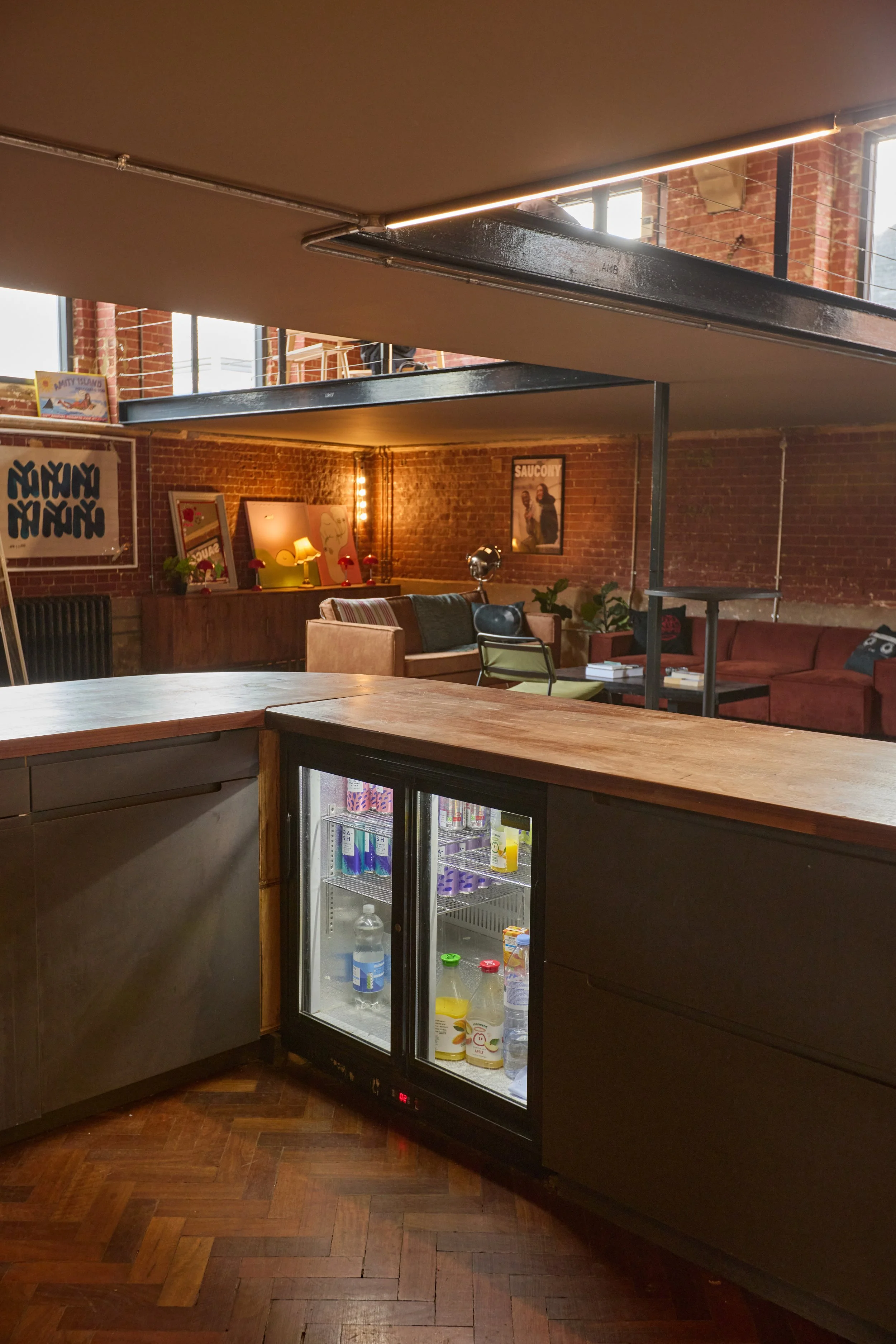 Interior of a lounge or cafe with exposed brick walls, a small refrigerator filled with beverages, and cozy seating areas with sofas and tables.