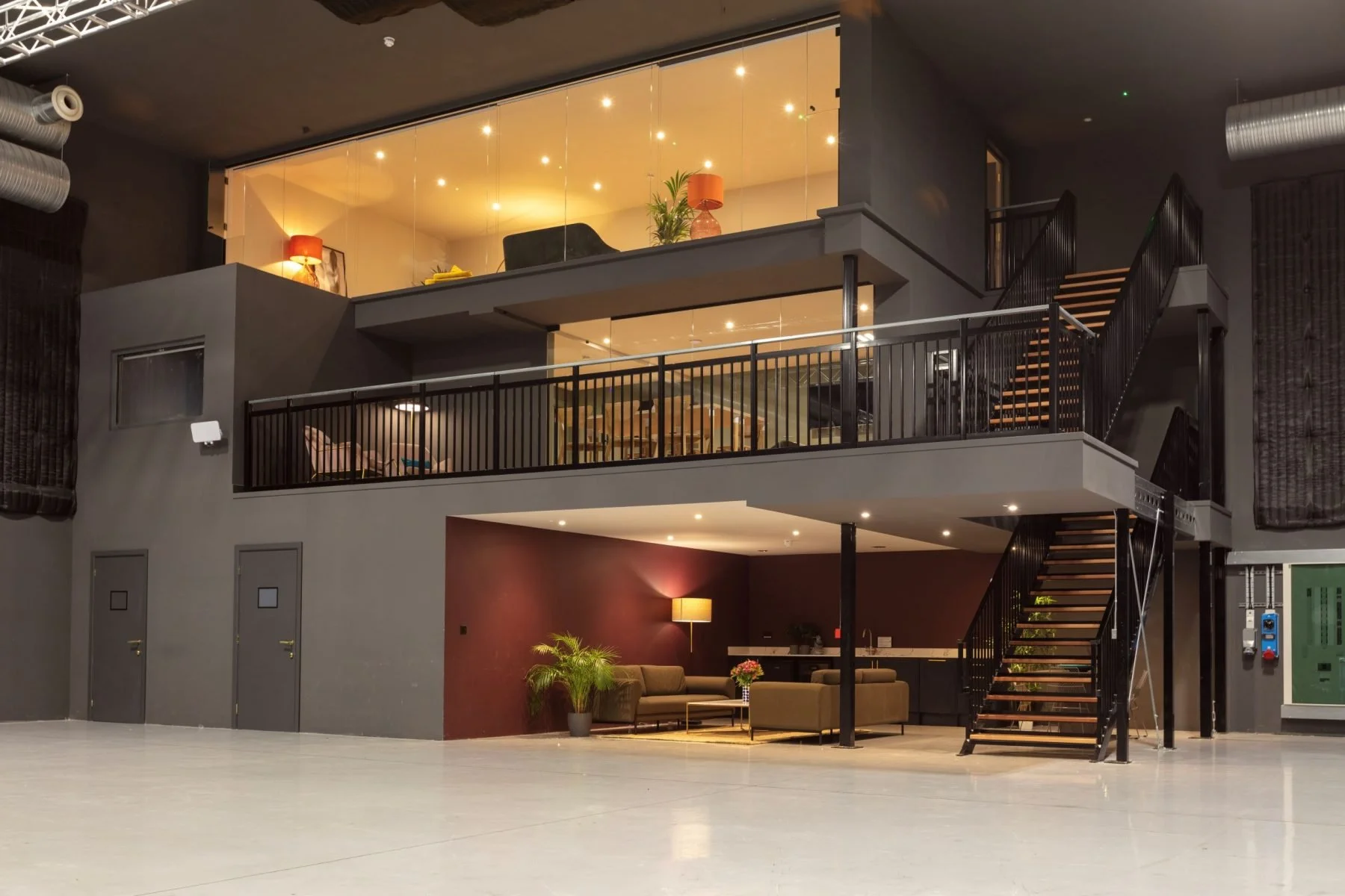 Interior of a modern, multi-story building with a ground floor lounge area featuring sofas, a lamp, and plants, and an upper level with tables, chairs, and decorative lamps, connected by black metal staircases.