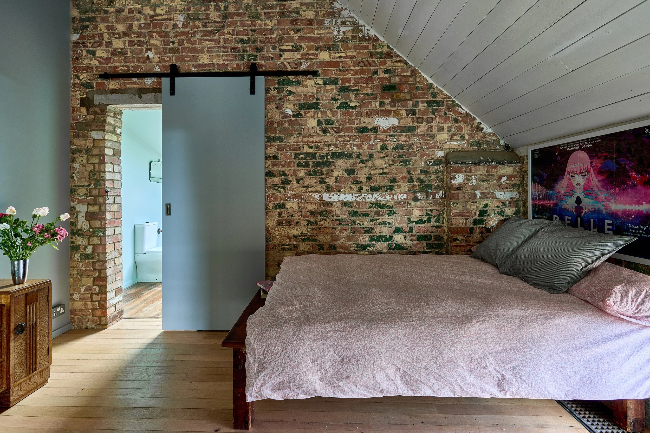 Bedroom with an exposed brick wall, a bed with pink bedding and gray pillows, a poster on the wall, a wooden side table with a vase of pink and white flowers, and a partially open sliding door leading to a bathroom with a toilet.