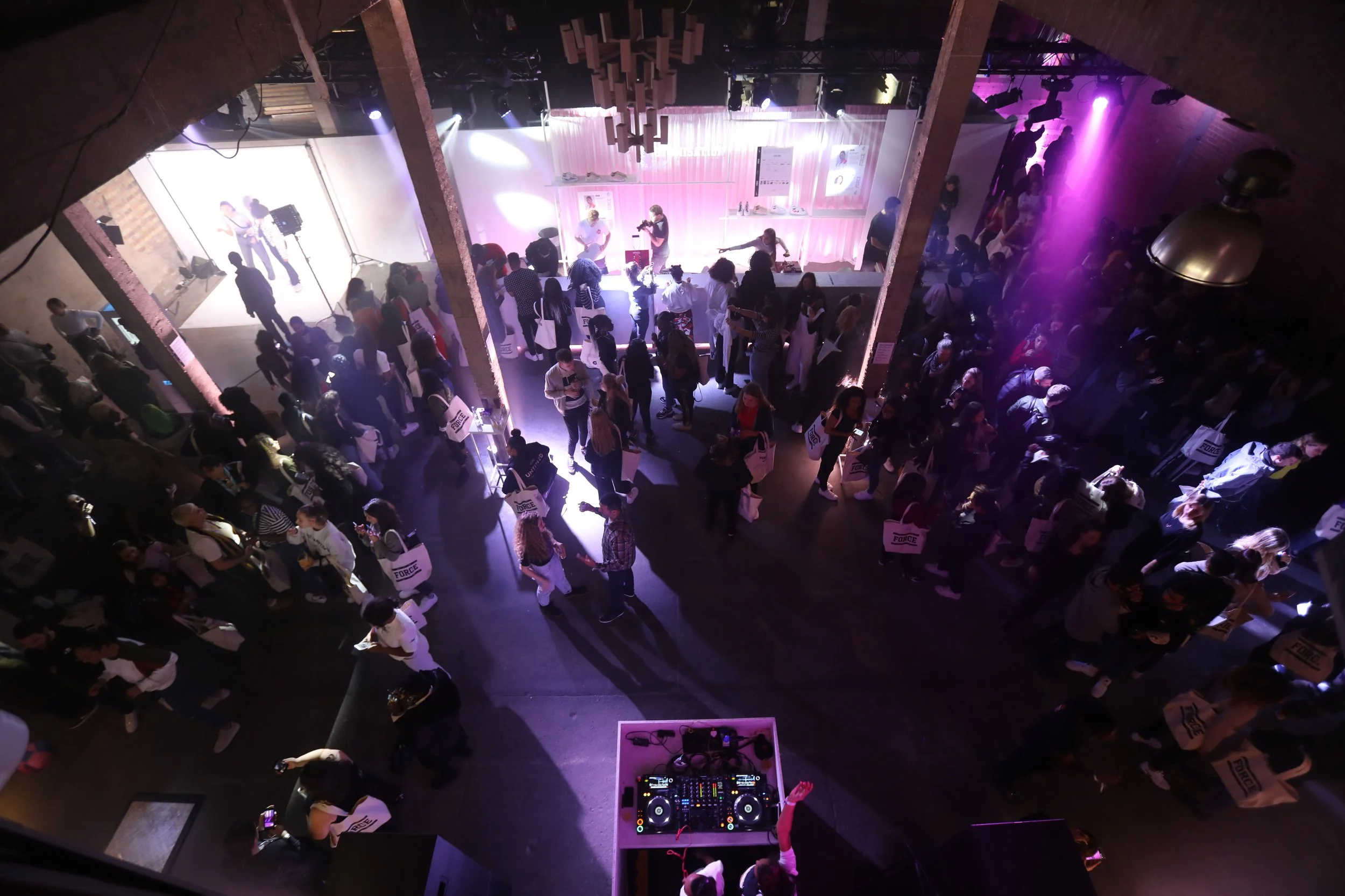 An indoor event with a DJ and dance floor, crowd of people standing and socializing, some dancing, with stage and purple lighting.