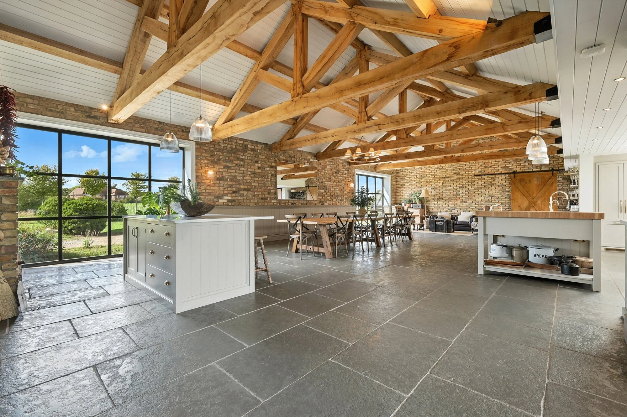 Open-plan kitchen and dining area with exposed wooden beams, brick walls, large windows showing a green outdoor landscape, and modern lighting fixtures.