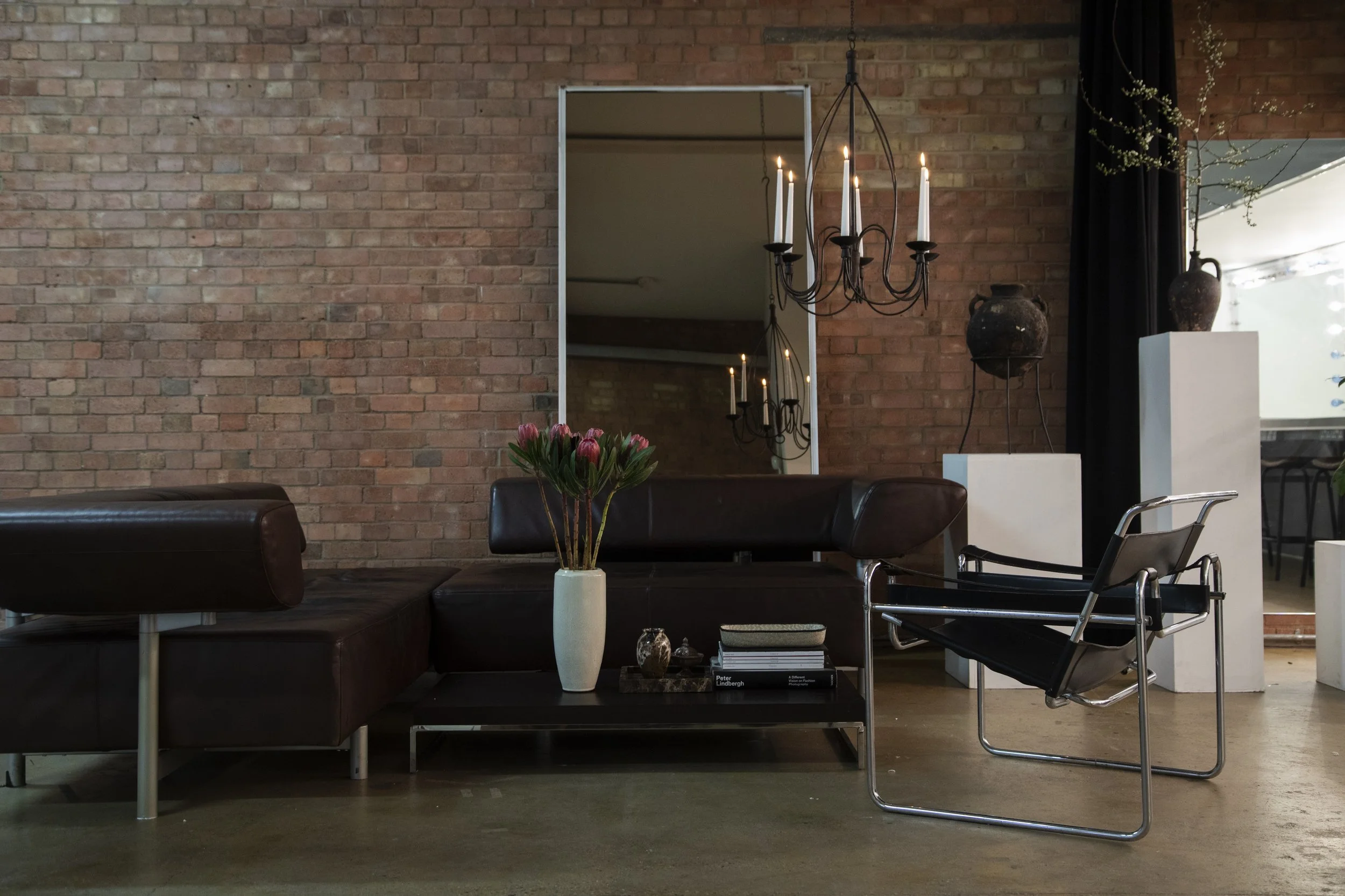 Living room with exposed brick wall, a large mirror, black leather sofa, modern chair, flower vase with pink tulips, books, a decorative box, a small sculpture, hanging chandelier with candles, and ceramic vases on white pedestals, with a window part