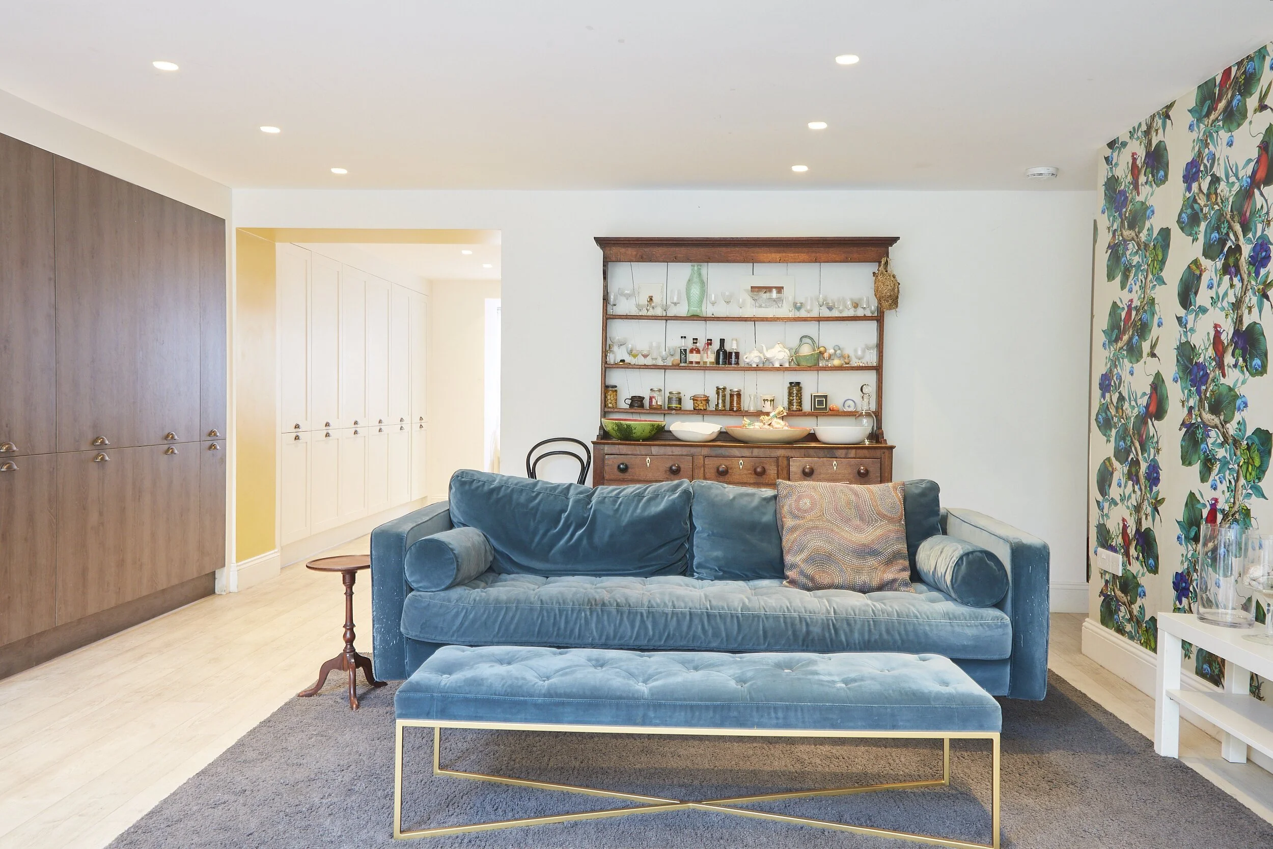 Living room with blue velvet sofa, wooden cabinet with shelves, and a floral accent wall.