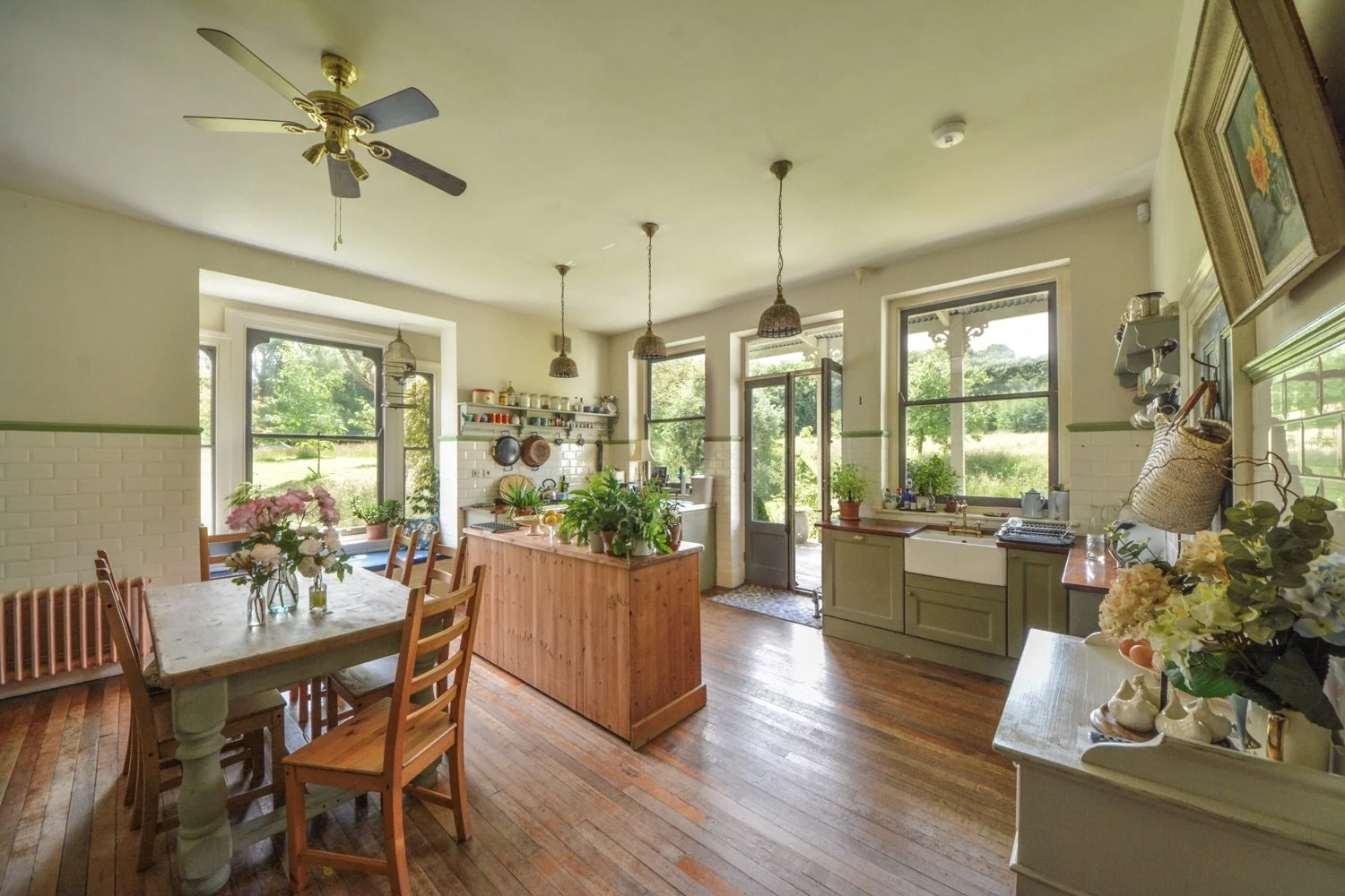 Bright kitchen and dining area with large windows, wooden dining table with chairs, hanging pendant lights, green cabinetry, and a central wooden island, decorated with plants and flowers.