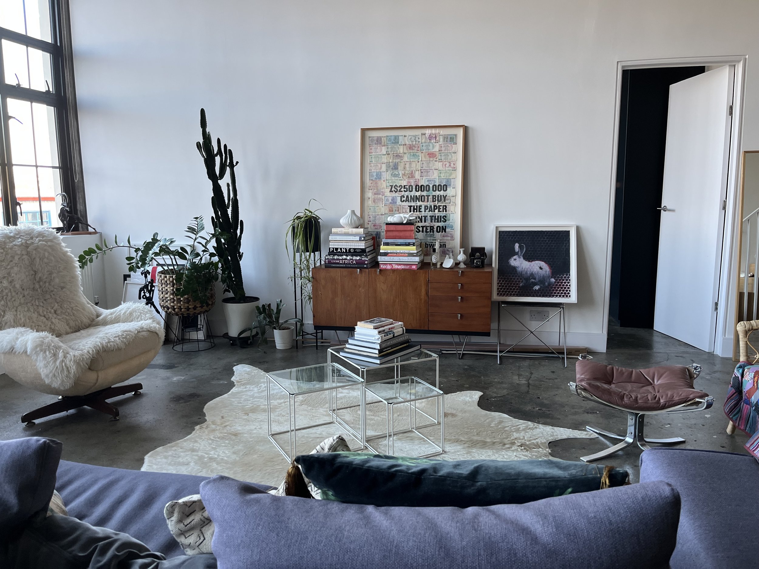 Living room with large window, white walls, and various indoor plants, including a tall cactus and a potted plant. There is a beige armchair with a fuzzy white throw, a modern glass coffee table, and a cowhide rug on the concrete floor. A sideboard w