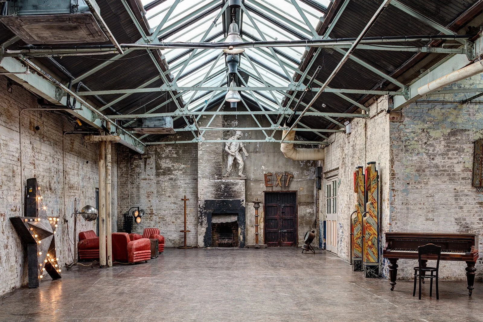 An industrial-style art gallery with exposed brick walls, metal beams, and a skylight ceiling. The room features vintage furniture, including red velvet chairs, a wooden piano, and colorful art pieces.