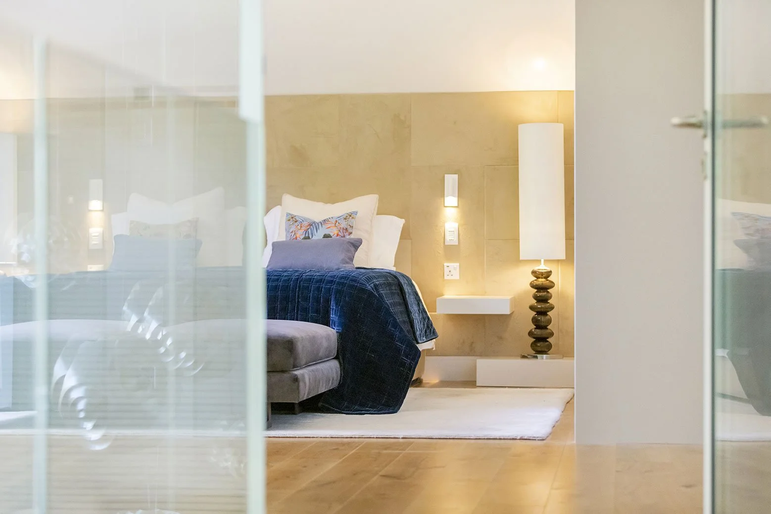 View of a modern bedroom with a bed featuring white and patterned pillows, a dark blue bedspread, a white side table, a tall white floor lamp, and wall-mounted lights, seen through glass panels.