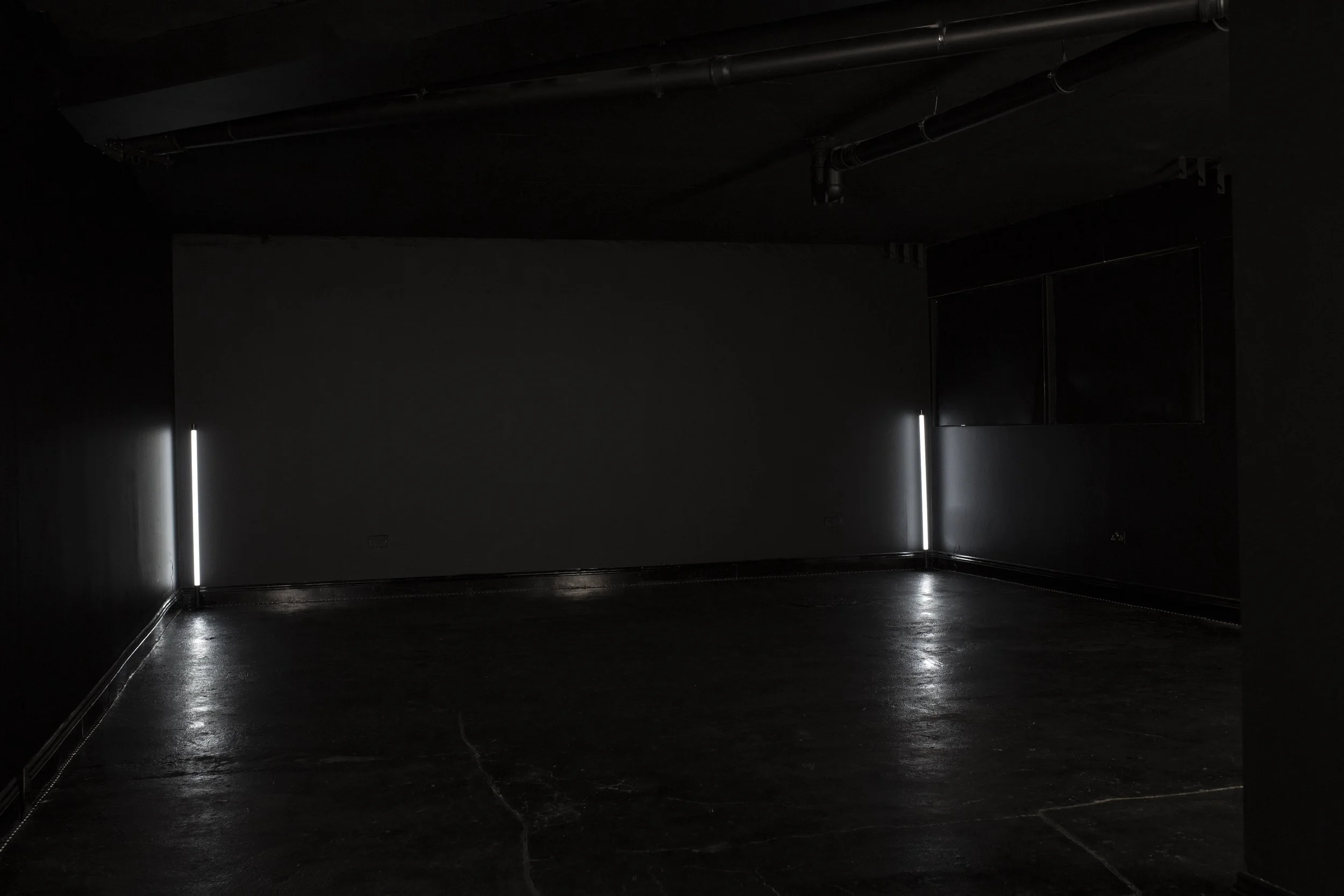 A dark room with black walls and floor, illuminated by vertical white light strips on two corners, with visible ceiling pipes.