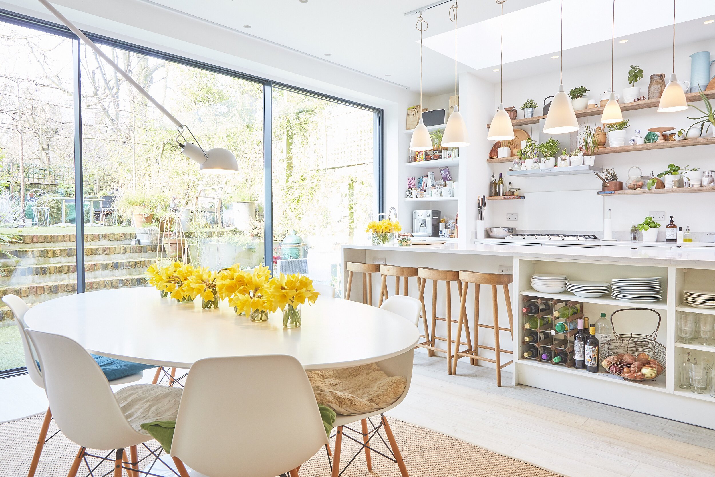 Bright, modern kitchen and dining area with large sliding glass doors leading to an outdoor patio. The room has white walls and cabinetry, open wooden shelves with plants and decor, a white kitchen island with bar stools, a round white dining table w