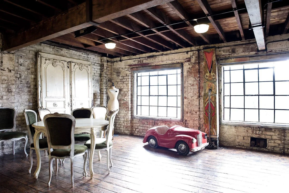 Interior of a rustic room with wooden floors and brick walls, featuring a vintage red toy car, a white ornate dining table with chairs, a mannequin, large industrial windows, and vintage decor.