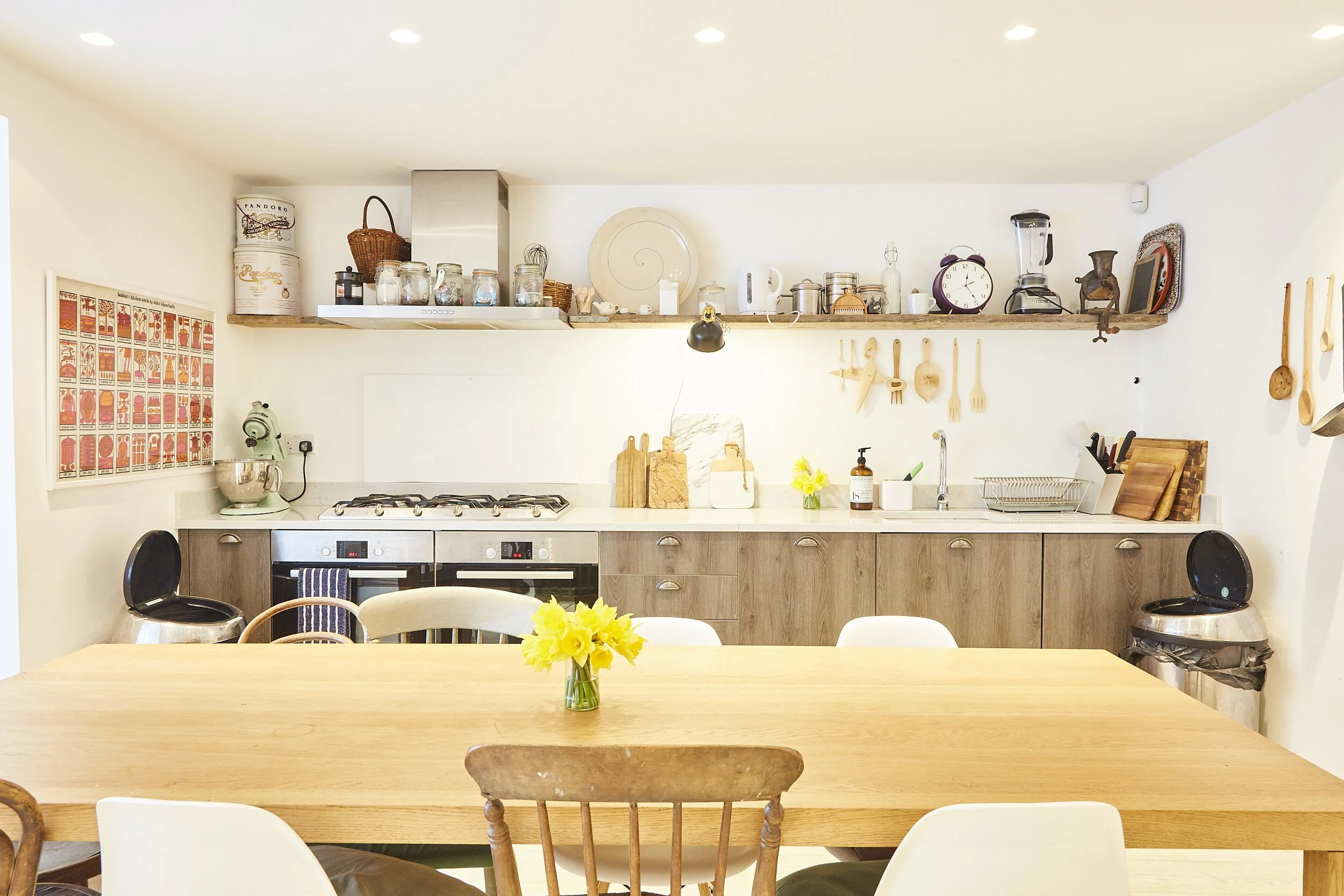 Bright, modern kitchen with wooden cabinets, open shelf with jars, kitchenware, and decor, dining table with yellow flowers in a vase, and various kitchen appliances and utensils.