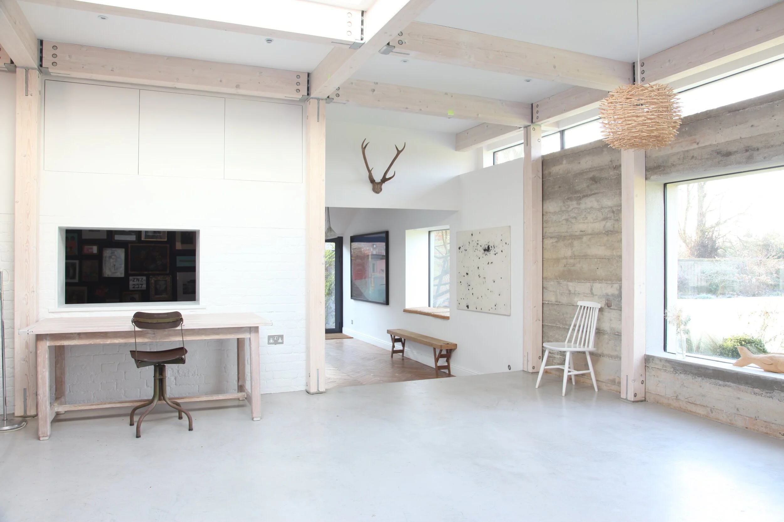 Open, modern interior with white walls, large windows, a desk with a chair, wall art, a deer antler mount, and minimal furniture.