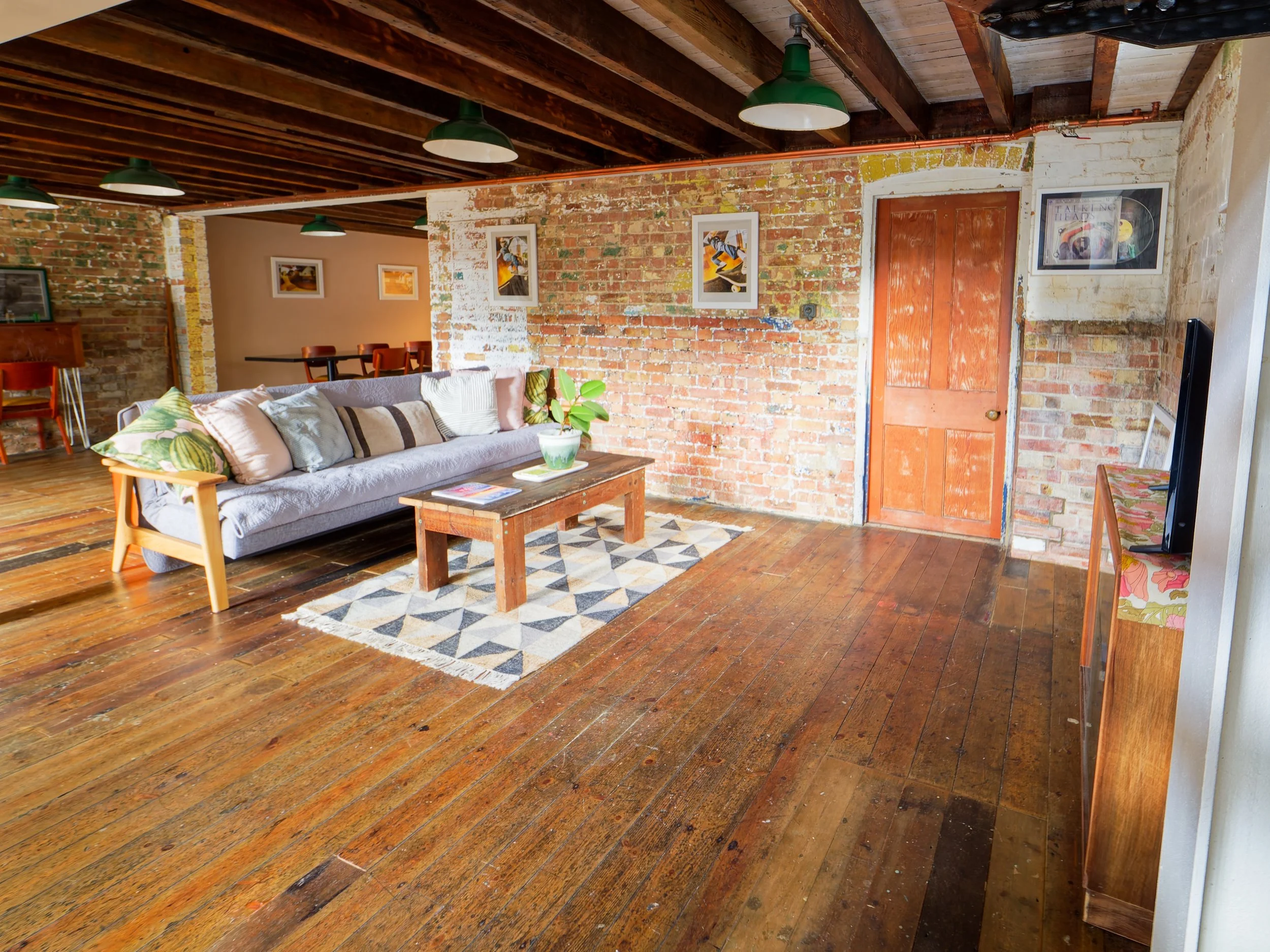 Living room with wooden floor, exposed brick walls, hanging green lamps, and a gray sofa with multiple pillows. There is a wooden coffee table with a plant and magazines, and a patterned rug. The door is wooden, and there is a TV on a stand to the ri
