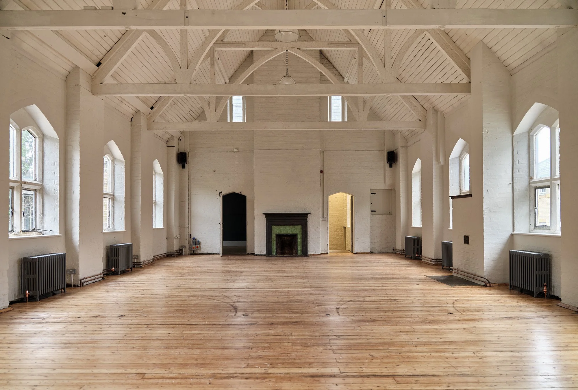 Empty room with white painted brick walls, wooden floor, high arched ceiling with white beams, multiple windows, a black fireplace, and radiators along the walls.