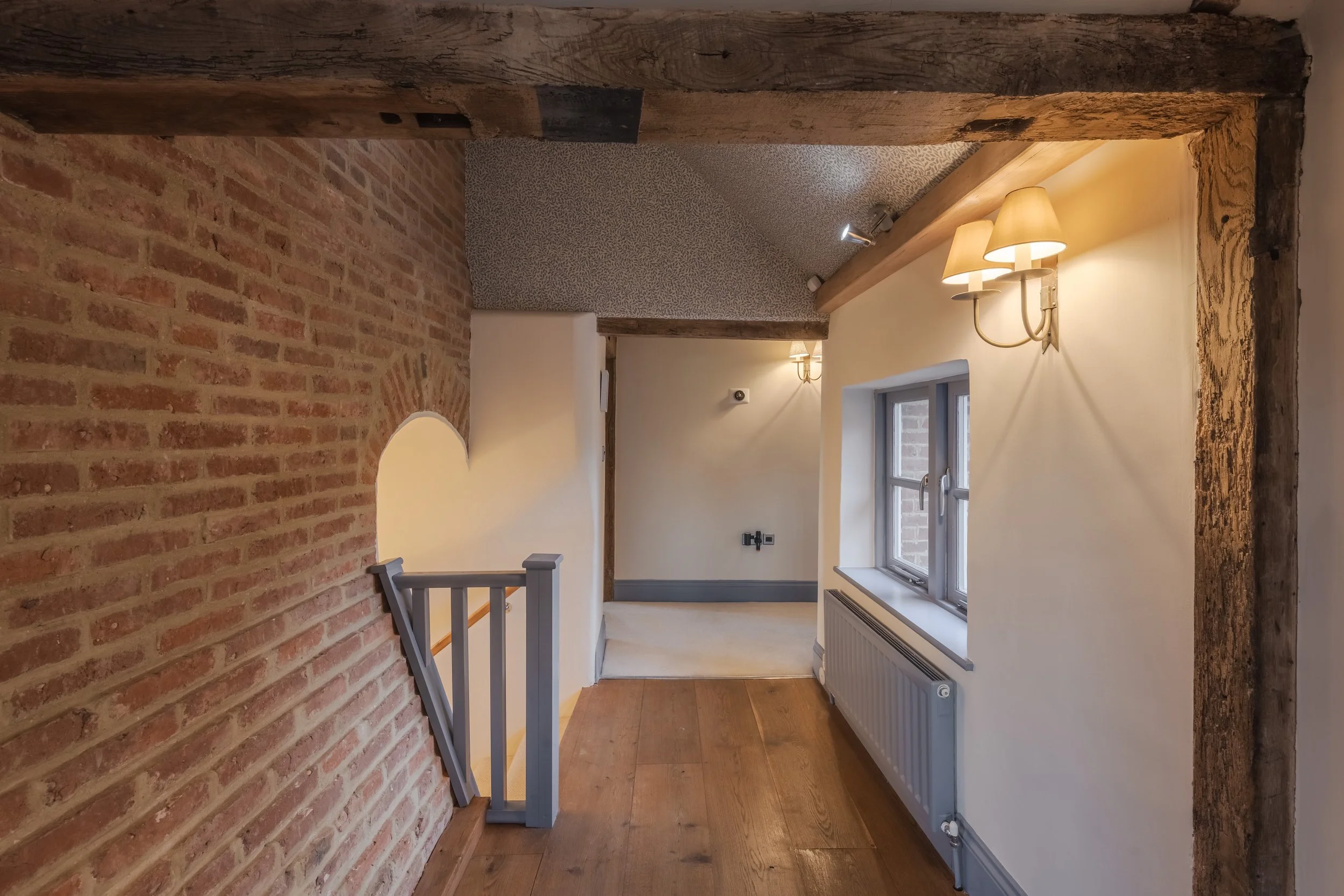 Interior view of a rustic-style room with exposed brick walls, wooden beams, a window, and warm wall-mounted lighting fixtures.