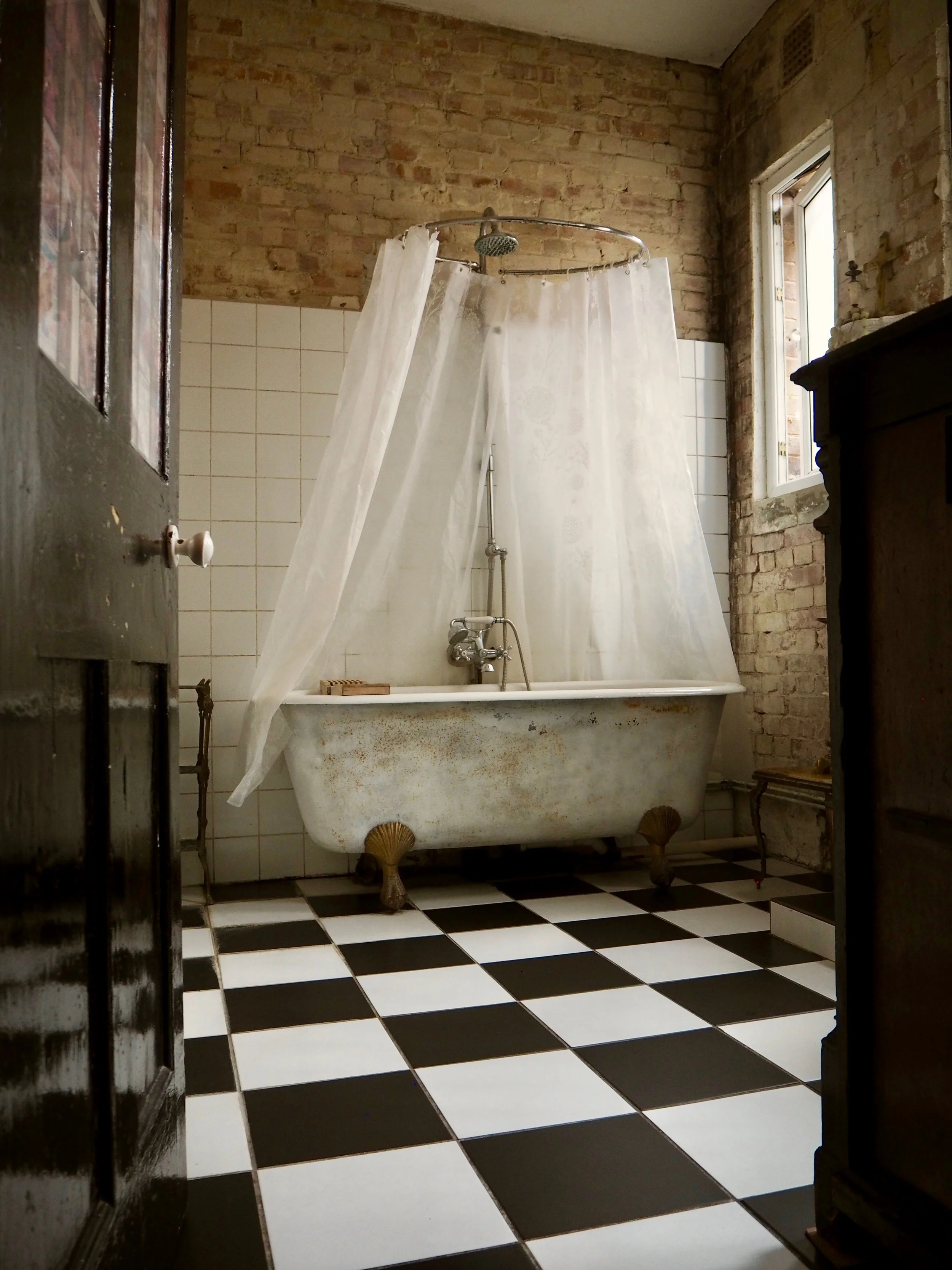 Vintage bathtub with clawfoot design in a rustic bathroom with checkered black and white tile floor, brick walls, and a shower curtain around the tub.