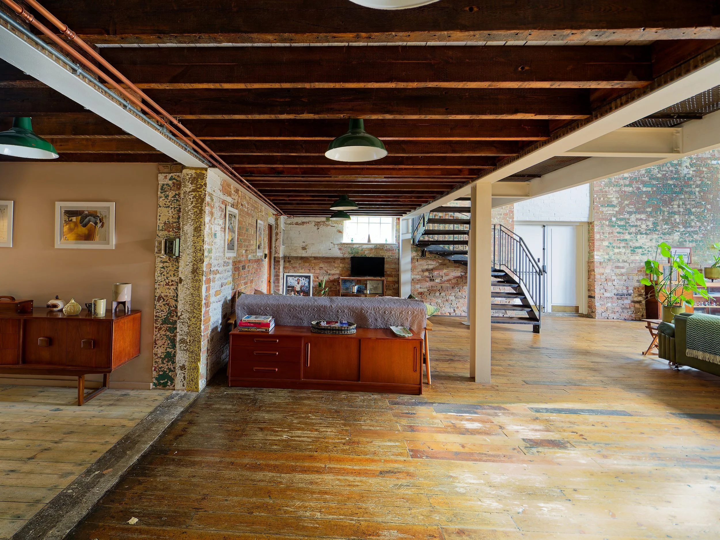 Interior of an industrial-style loft with exposed brick walls, wooden floors, and a mix of vintage and modern furniture including a brown sofa, side tables, and potted plants, with stairs leading to an upper level.