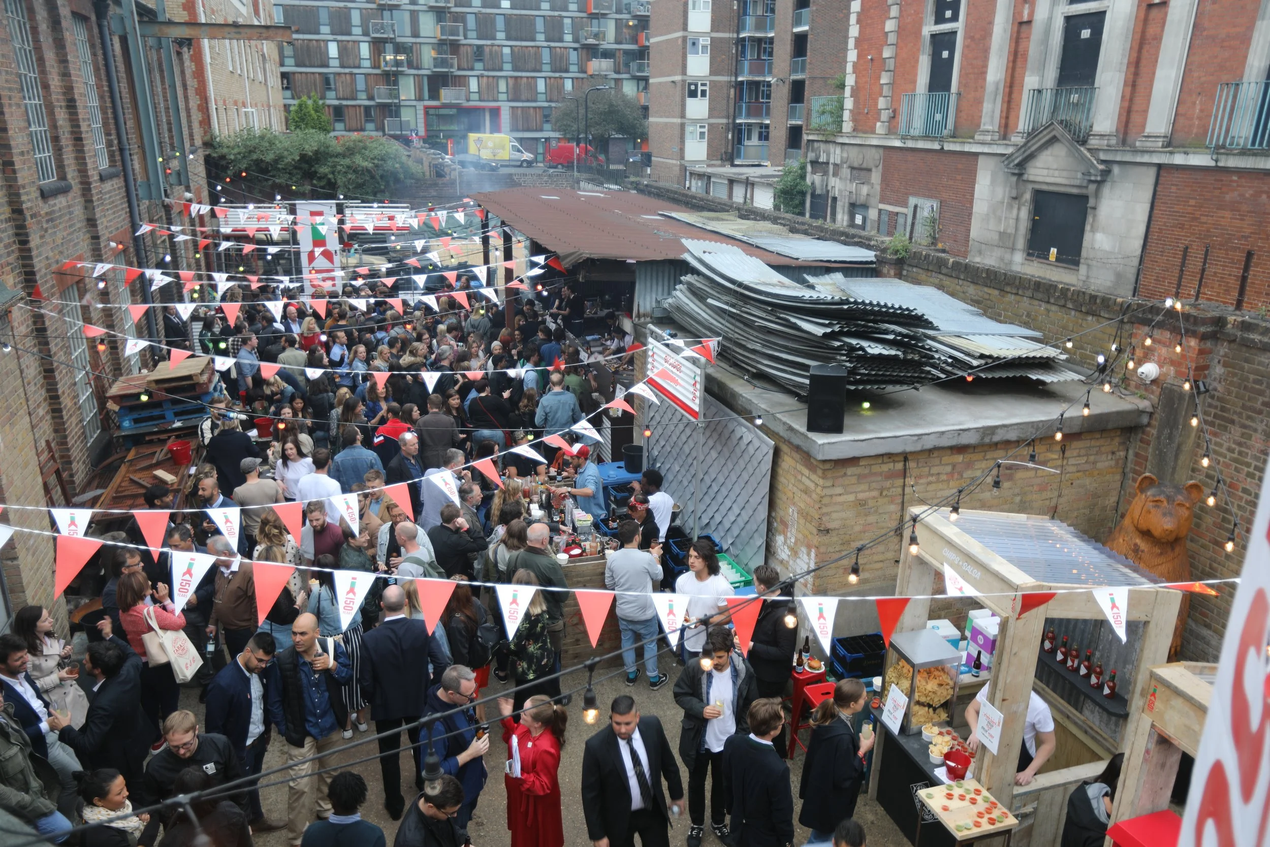 A crowded outdoor event or party with many people gathered under string lights and red and white bunting decorations, situated between brick buildings, with food and drink stalls visible.