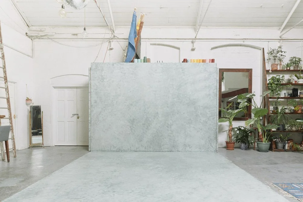 Interior of a creative workspace with a large light blue textured wall, potted plants, a wooden shelf with plants, a mirror leaning against the wall, and various art supplies on top of the wall, with white painted brick ceiling and concrete floor.