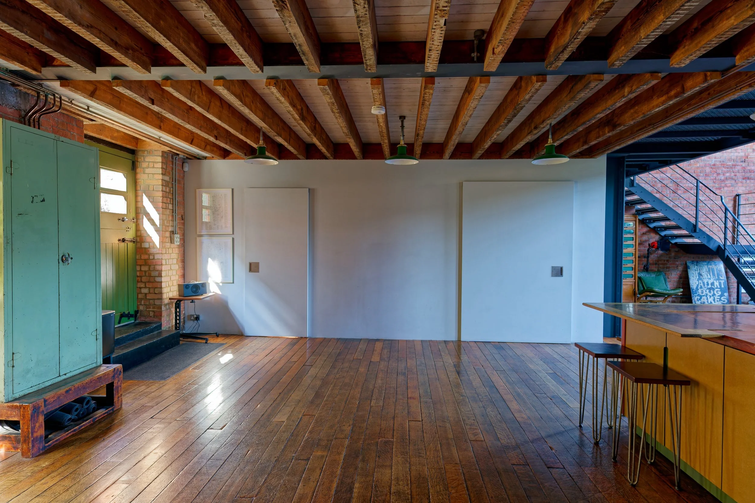 Empty industrial-style room with wooden floors, exposed wooden beams on the ceiling, a light green storage cabinet on the left, a small speaker on a stand, and a brick wall with three small windows. To the right, there are stairs leading up and a hig