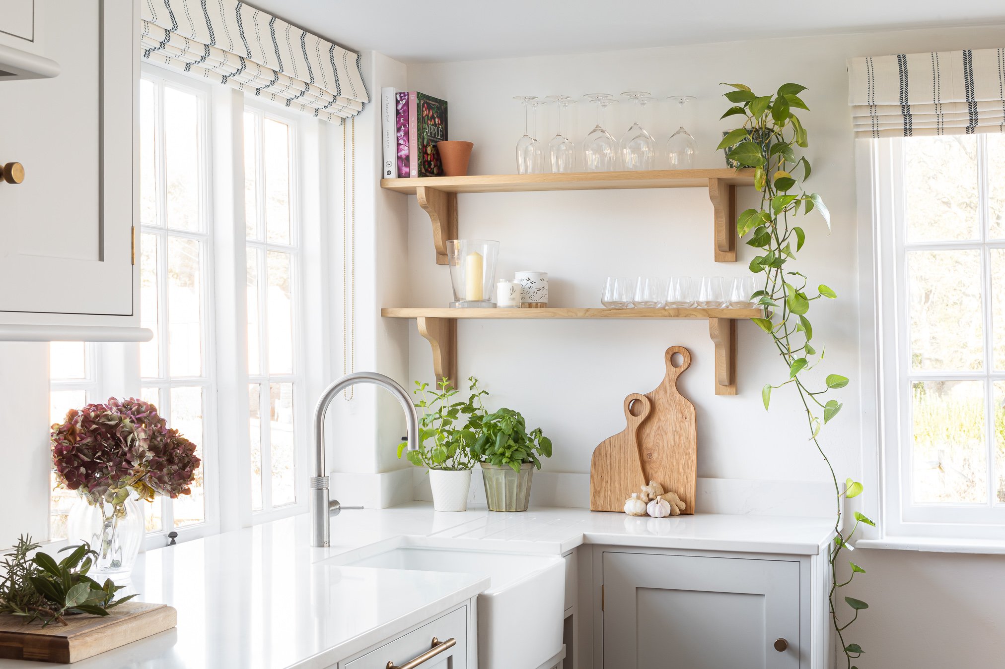 Bright kitchen corner with white cabinets, a white countertop, large windows with striped valances, and wooden shelves holding glassware, books, and plants.
