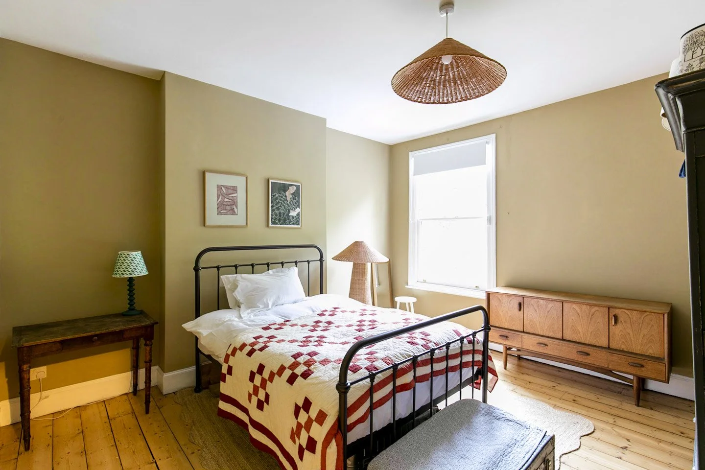 A bedroom with a black metal bed frame, a white quilt with red geometric patterns, and white pillows. There is a small wooden bedside table with a green and white patterned lamp on the left, a woven lampshade, a window with sunlight coming through, a