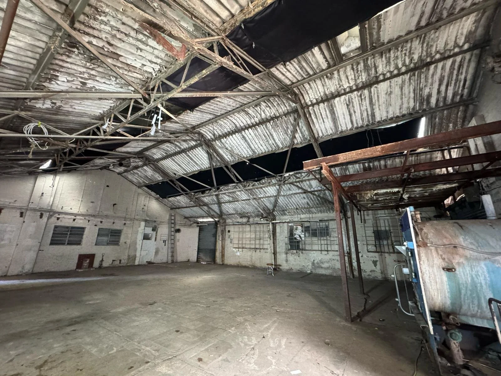 An empty, abandoned industrial warehouse with a damaged, rusted metal roof and peeling paint on the walls.