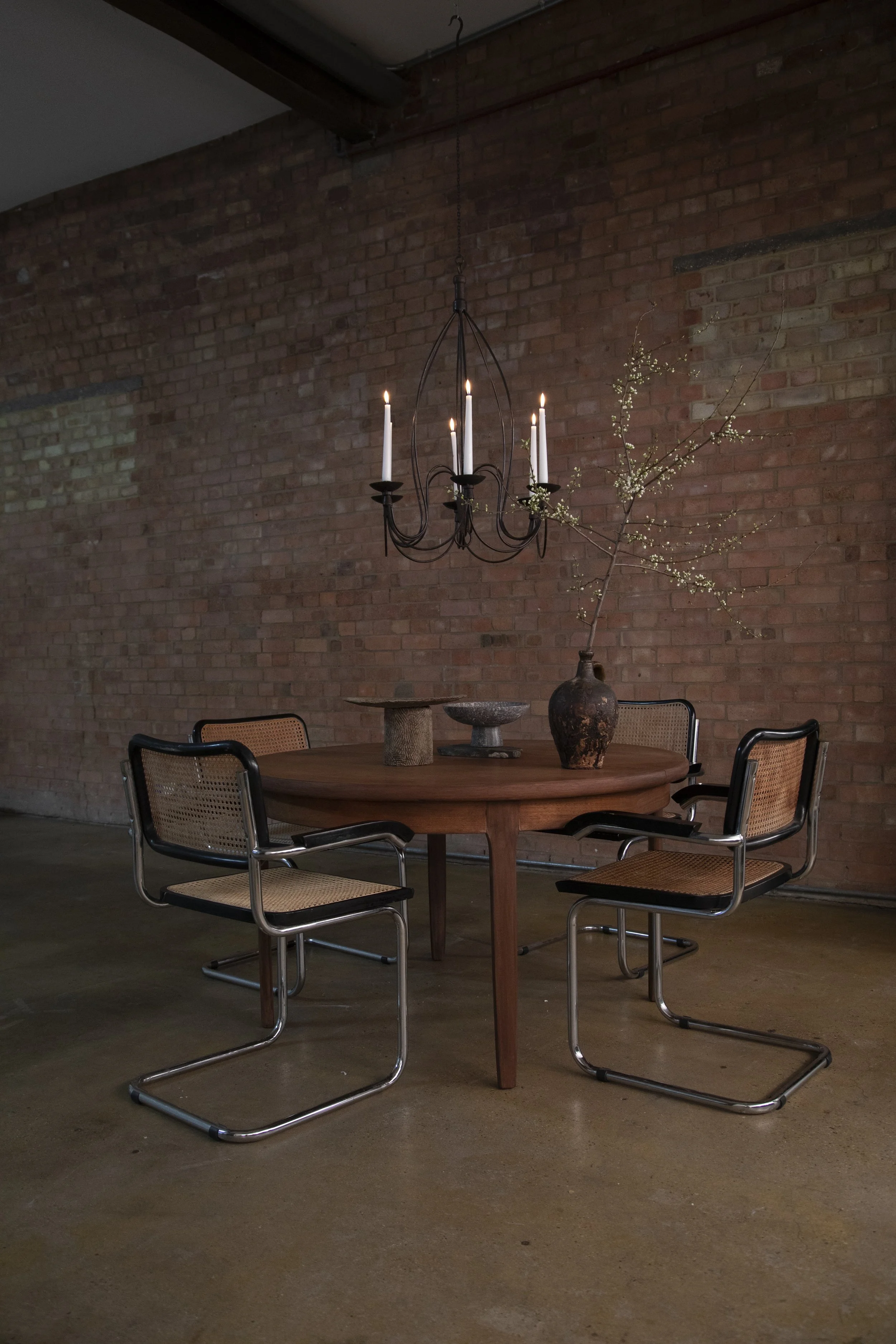 A round wooden dining table with three metal and cane chairs, a black wrought iron chandelier with lit candles hanging above, and a vase with branches and small flowers on the table. Brick wall background.