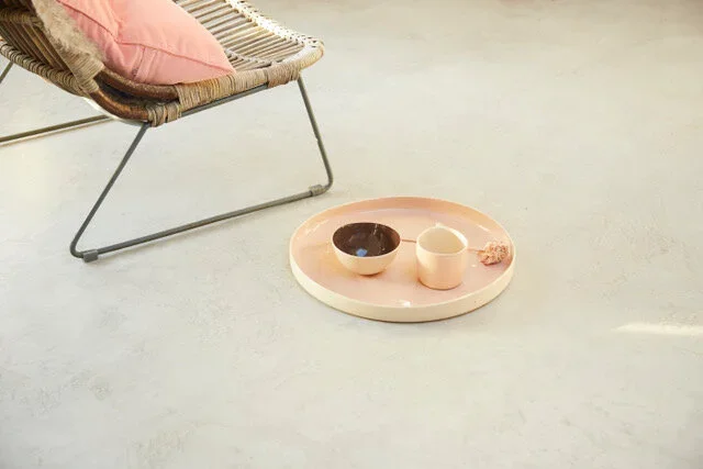 A pink tray with two cups and a small item on a concrete surface near a rattan lounge chair with a pink cushion.