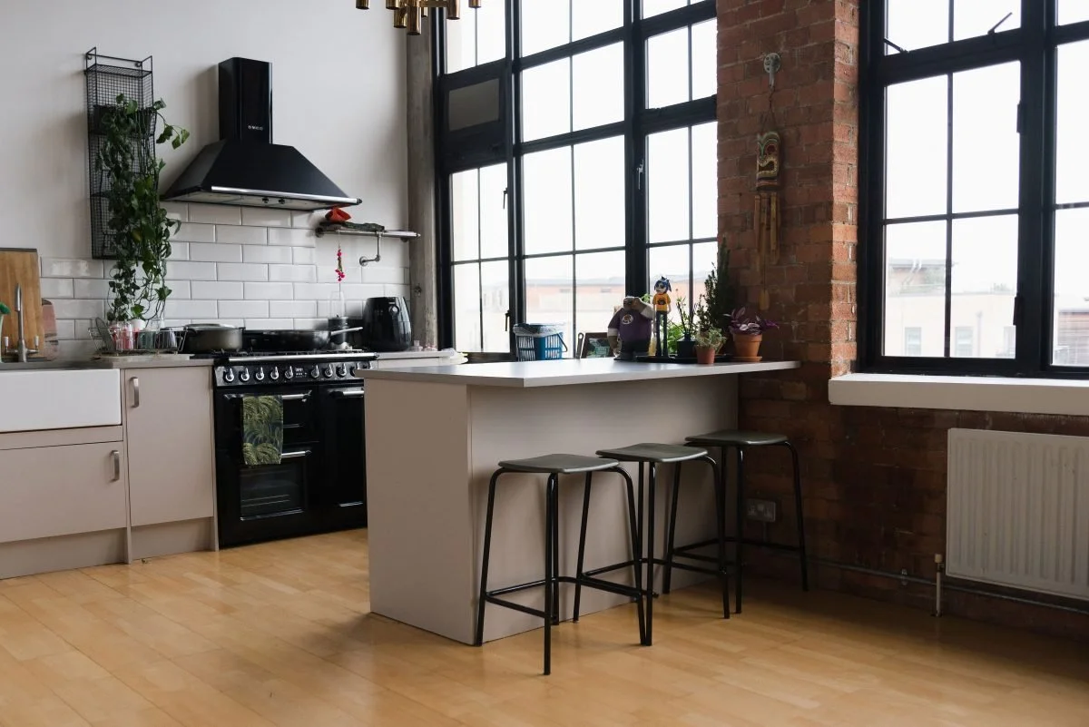 Modern kitchen with large windows, brick wall, white cabinetry, black stove, range hood, and a white island with three black stools.