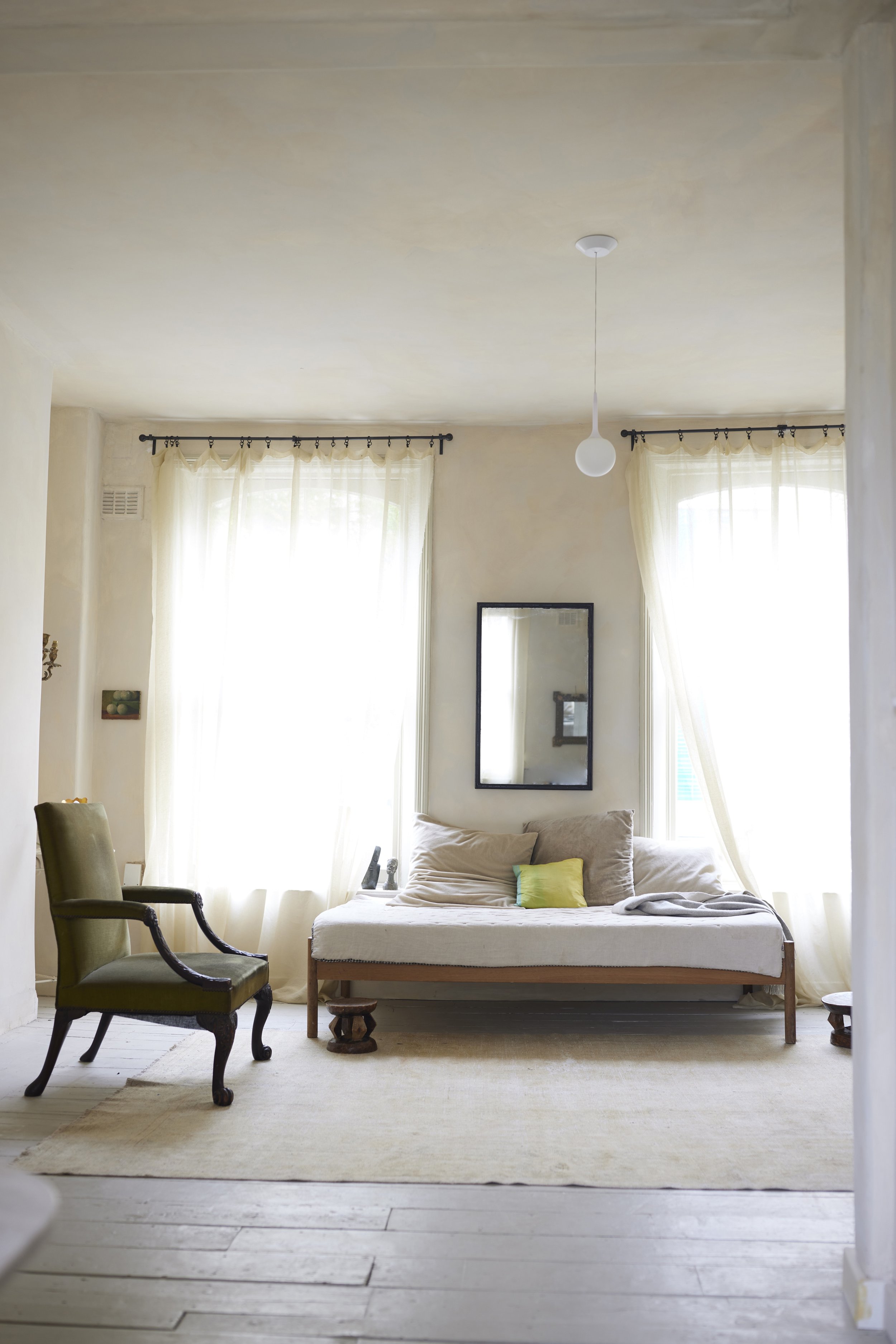 Bright living room with large windows, white curtains, a mirror on the wall, a daybed with pillows, an upholstered armchair, and a hanging ceiling light.