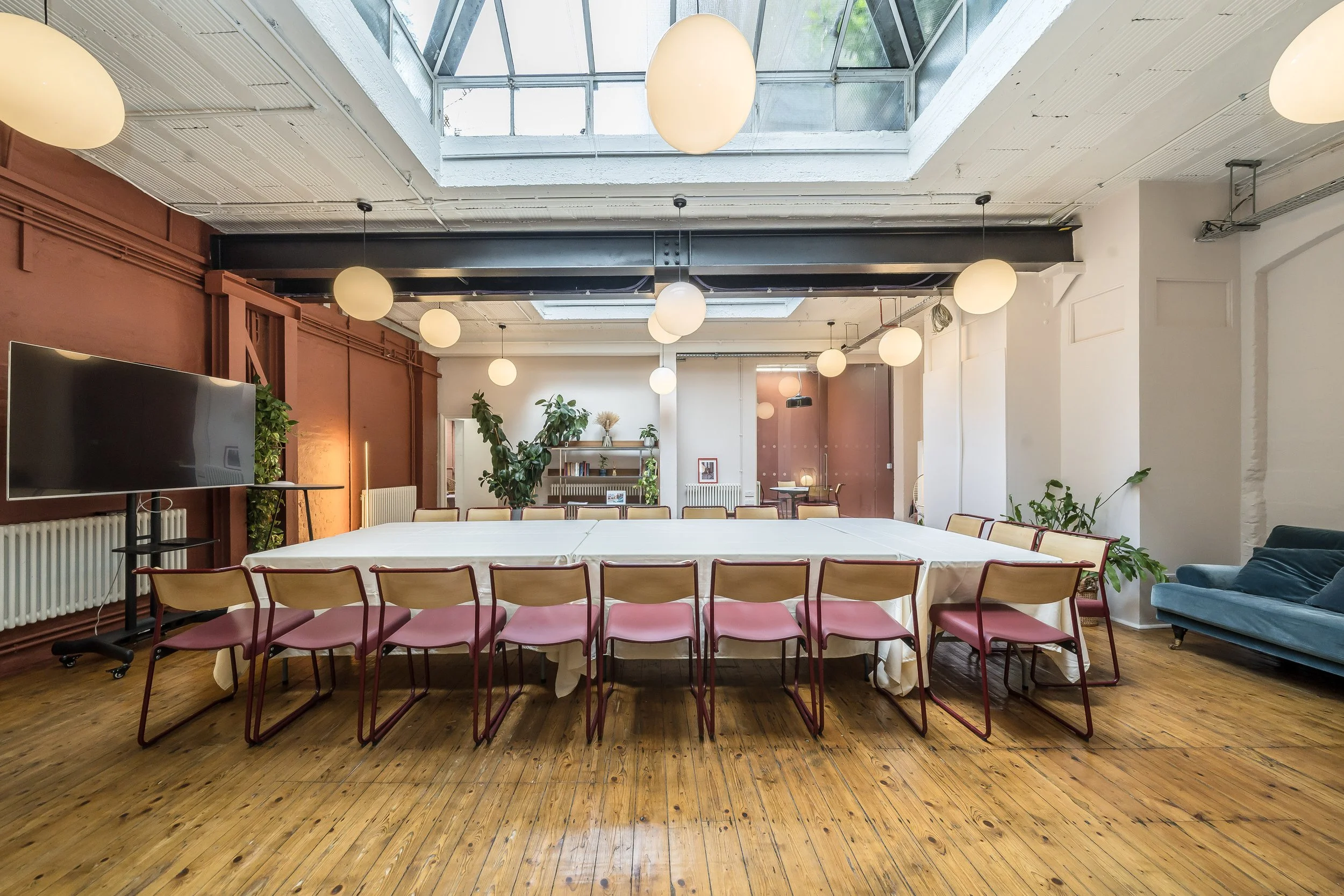 Large room with a long, rectangular table covered with a white tablecloth, surrounded by pink and beige chairs, wooden floor, plants, bookshelf, large windows, white and brown walls, ceiling with hanging round white lights, a TV on a stand, and a tea