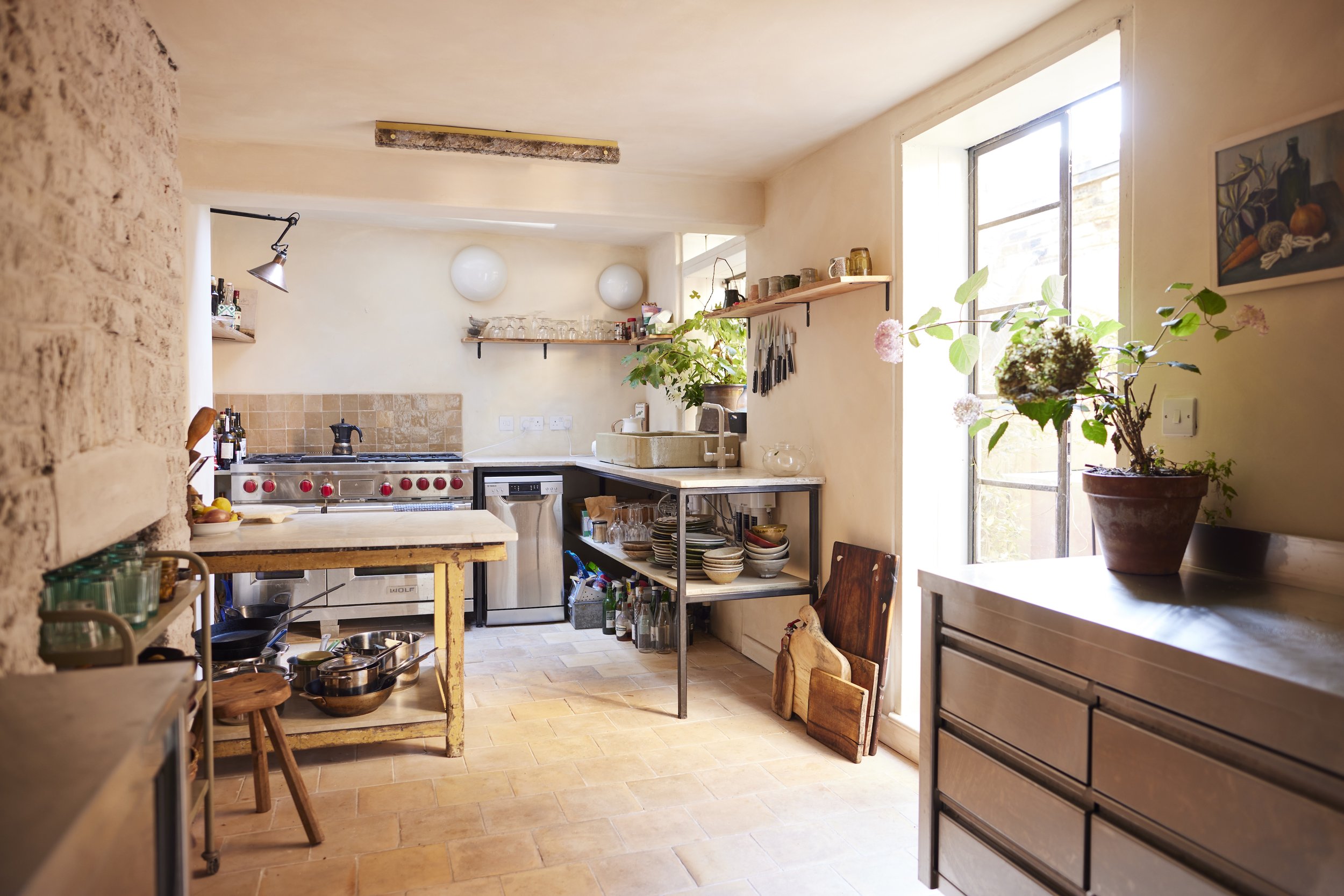 A cozy, rustic kitchen with a stone wall, wooden furniture, open shelving, a window with sunlight, various kitchenware, potted plants, and a beige tiled floor.