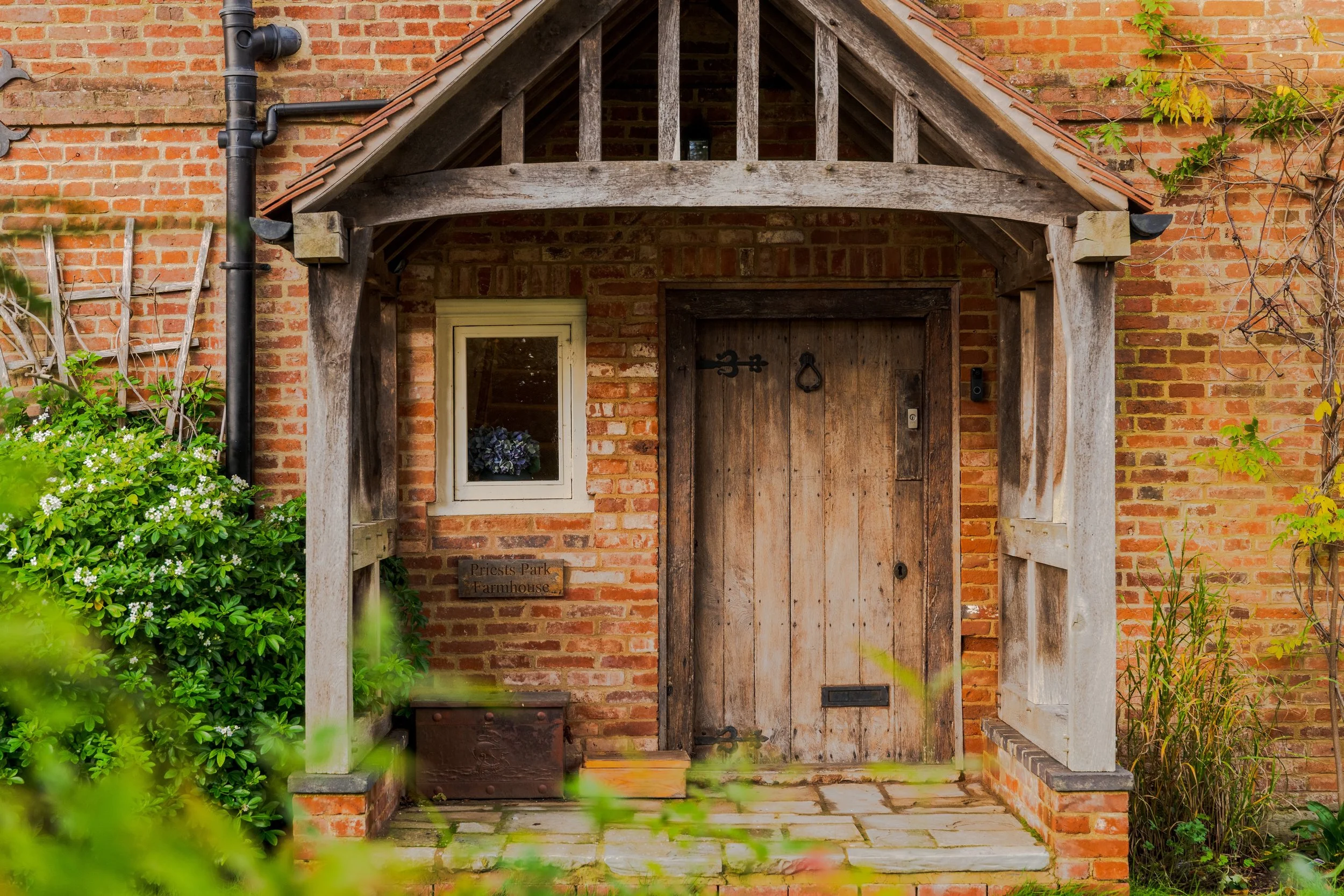 A rustic farmhouse entrance with a weathered wooden door, small window, brick walls, and a wooden porch structure. Green foliage surrounds the entrance.