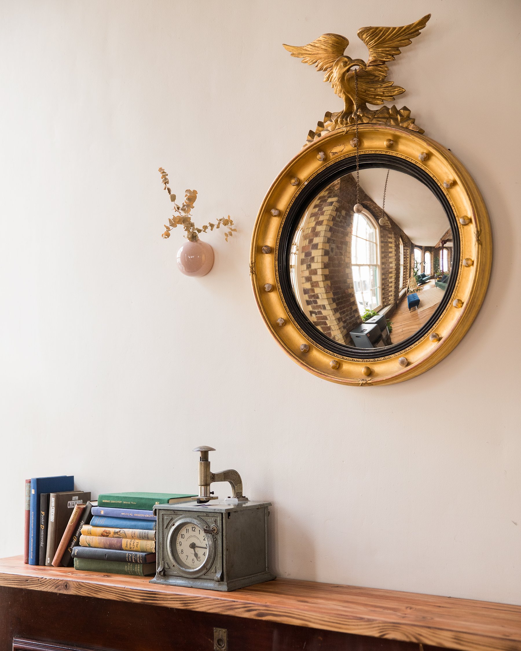 Interior decor with vintage elements including a round gold-framed mirror, a brass eagle sculpture on top of the mirror, a pink vase with dried plant, a stack of books, a vintage metal desk clock, and a reflection of a brick wall and windows in the m