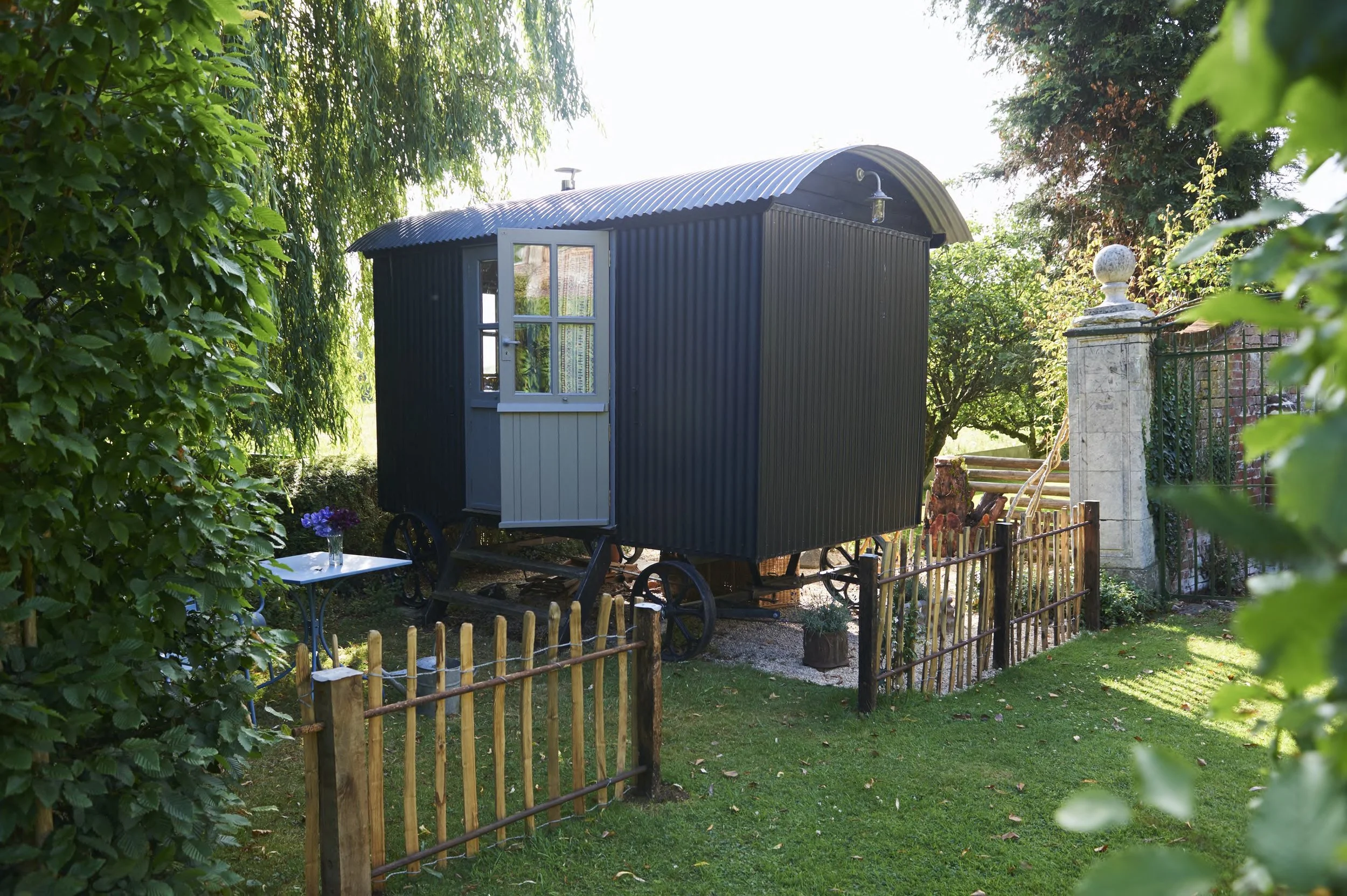 A black tiny house or shed on wheels with a door and window, situated in a garden with greenery and a small fence.
