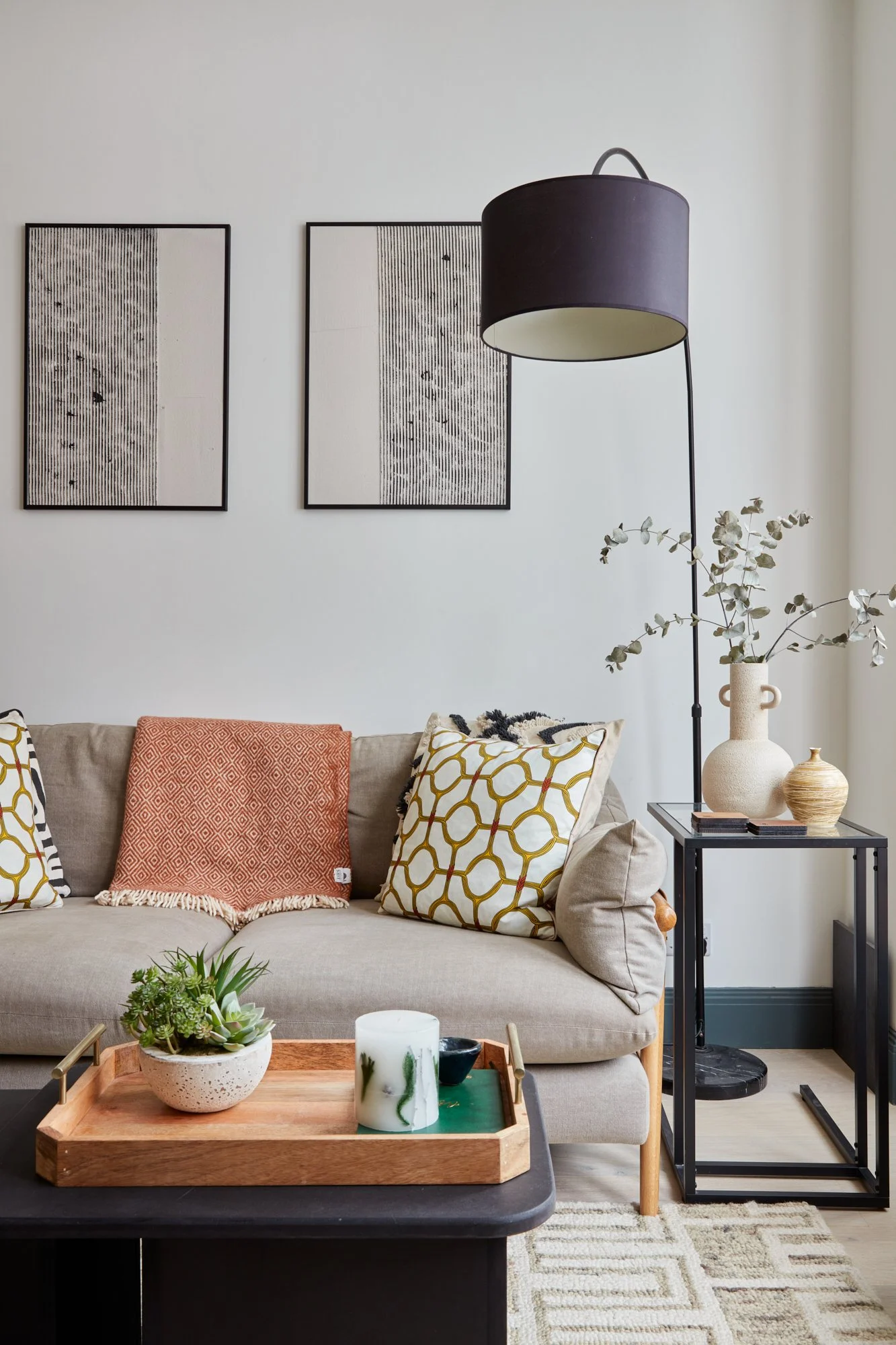 Living room with beige sofa, patterned pillows, black side table with vases and a tall black floor lamp, wall art, and a wooden tray with plants and candles.