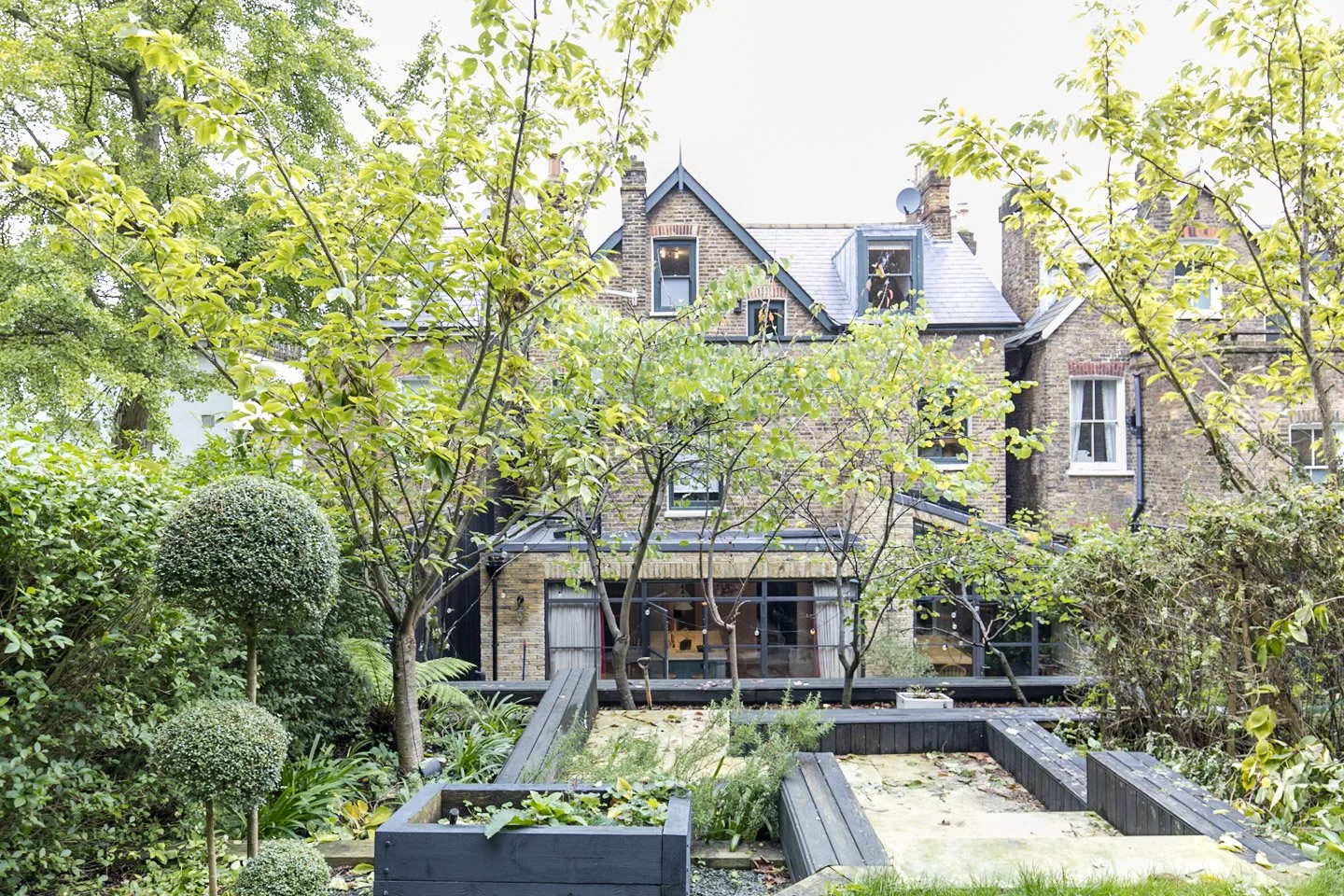 Backyard garden with trees, shrubs, and a brick house with multiple chimneys and windows.