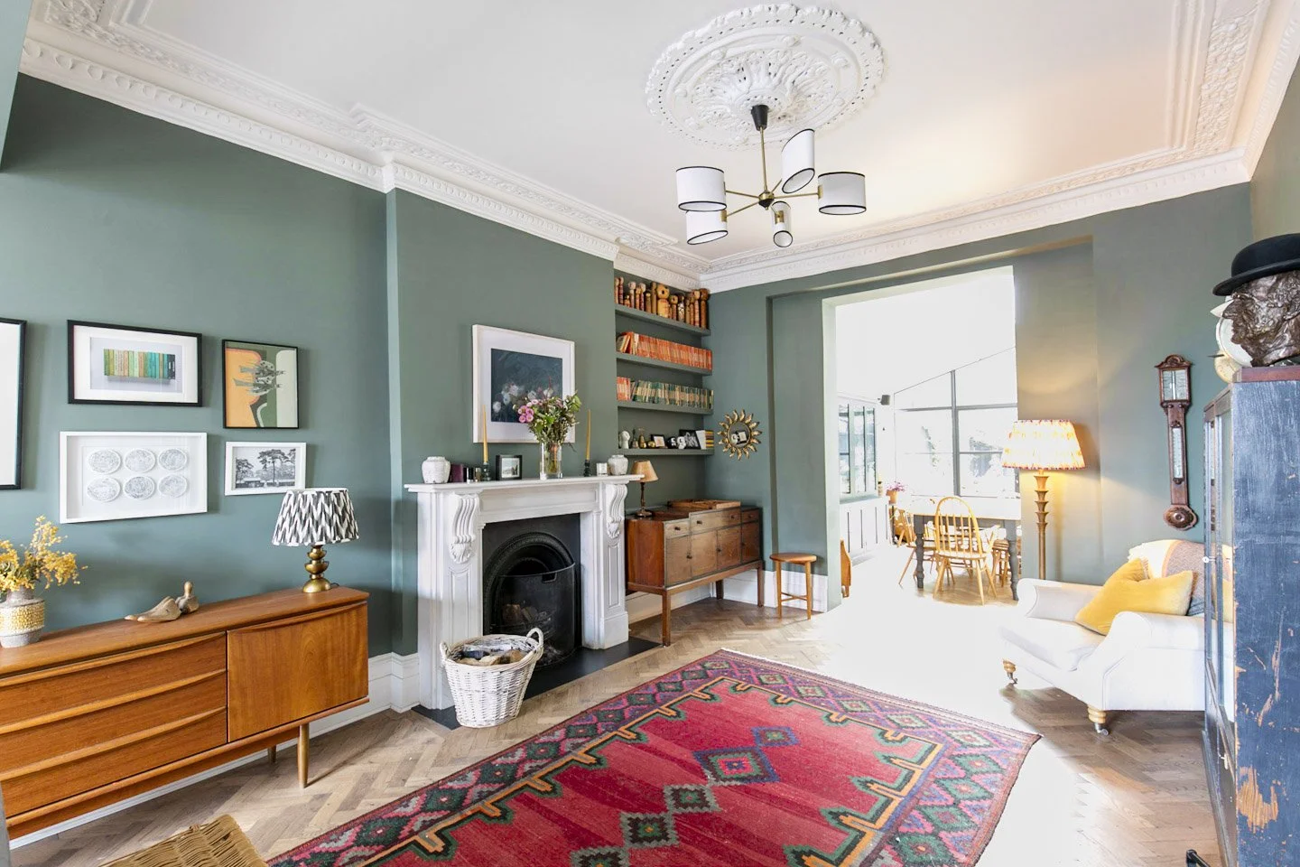 Living room with green walls, decorative ceiling molding, artwork, a white fireplace with a floral arrangement, wooden furniture, patterned rug, a white couch, and a sunlit dining area in the background.