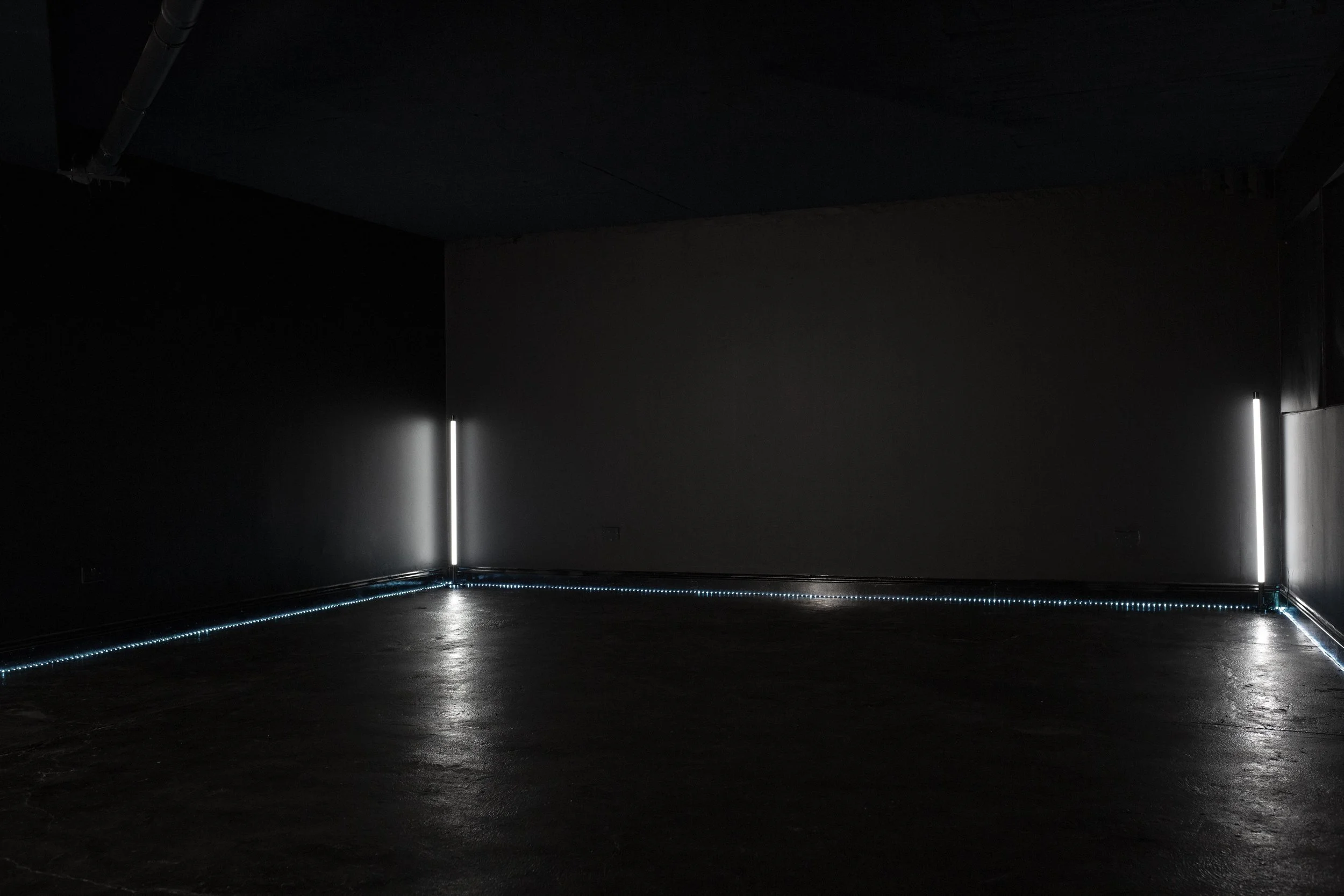 Dark room with minimal lighting, black walls, floor, and ceiling. Vertical white light strips on the left and right walls, and a blue LED strip along the floor.